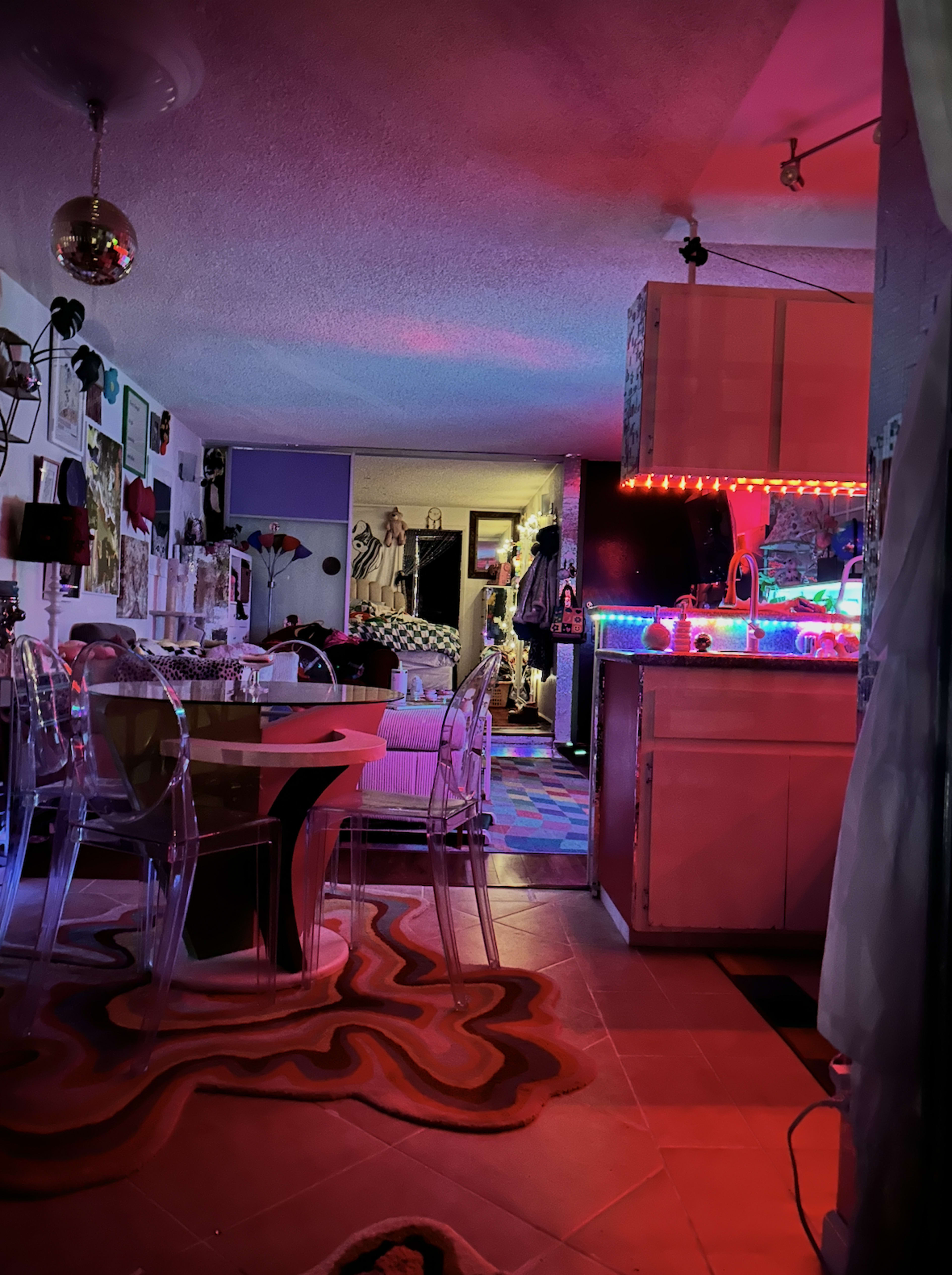 The image shows a colorful, eclectic living space with bright lighting, a dining area with transparent chairs, and a kitchen featuring multicolored LED lights.