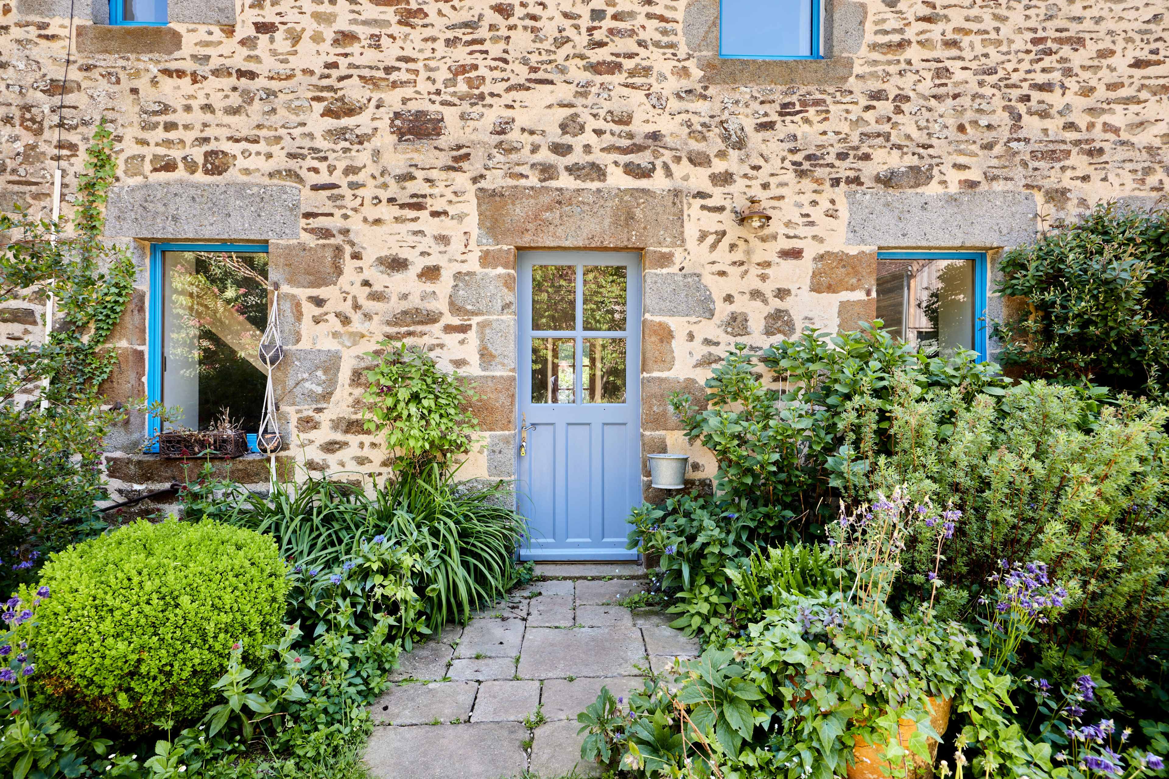 The image depicts a stone cottage with a blue door surrounded by greenery and flowers in a garden.