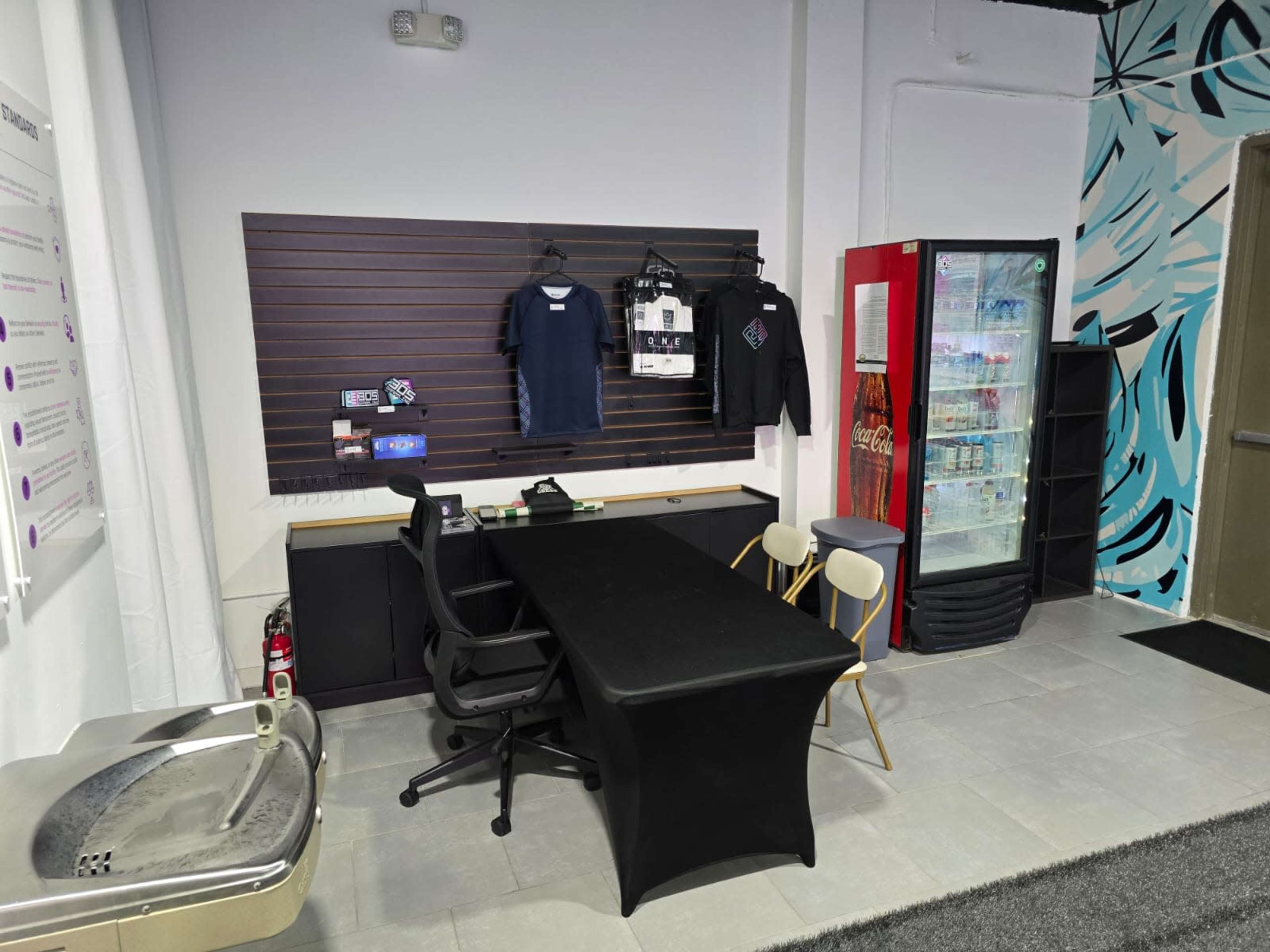 The image shows a small office area featuring a black table and chairs, a display wall with clothing, and a beverage cooler next to a water fountain.