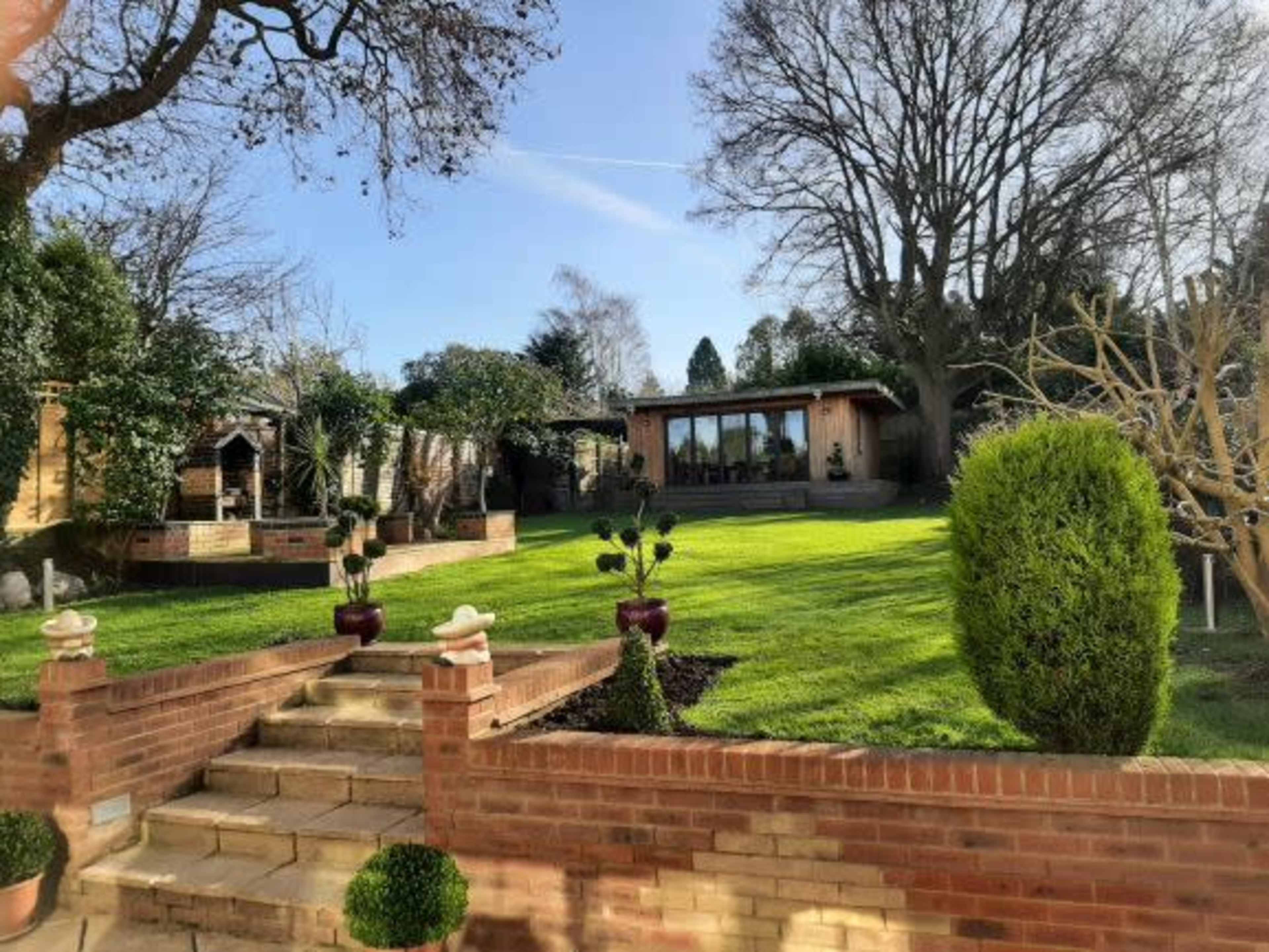 The image shows a landscaped garden with a stone path leading up to a wooden cabin surrounded by trees and neatly trimmed bushes.