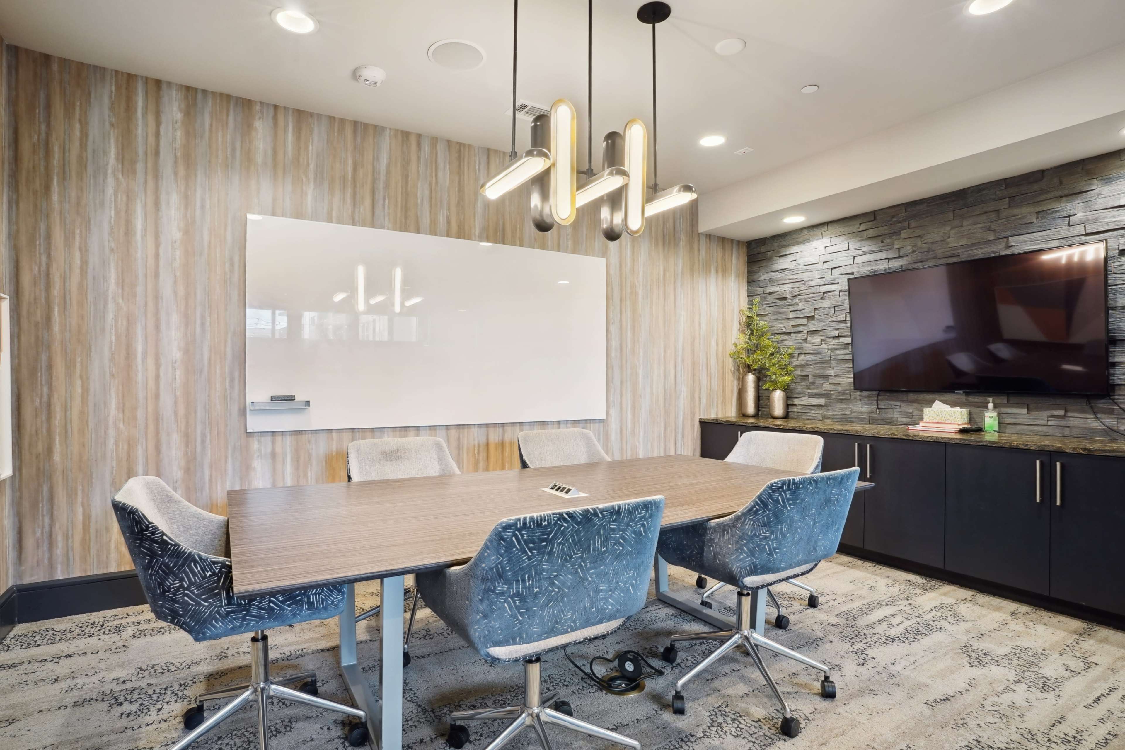 A modern conference room features a long wooden table surrounded by gray upholstered chairs, a whiteboard, a television screen, and a stone accent wall.