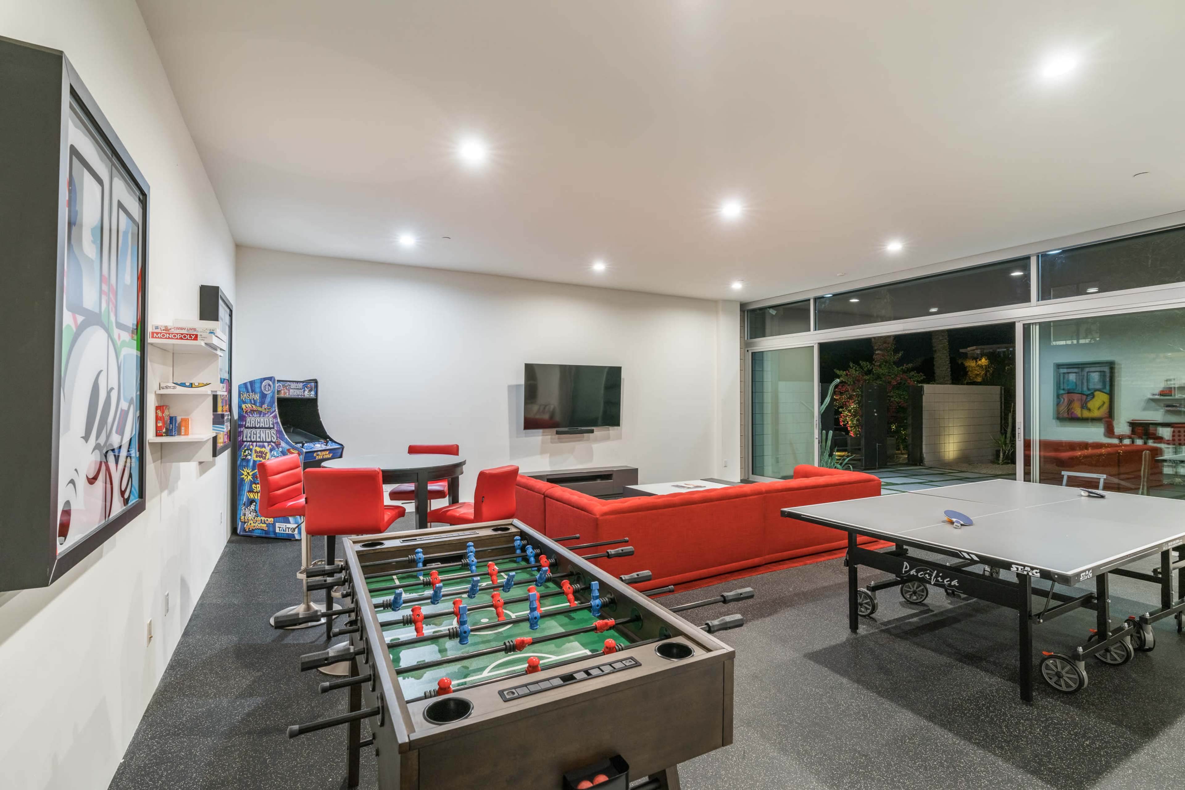 A modern game room featuring a foosball table, a ping pong table, a red sofa, an arcade machine, and a large television mounted on the wall.