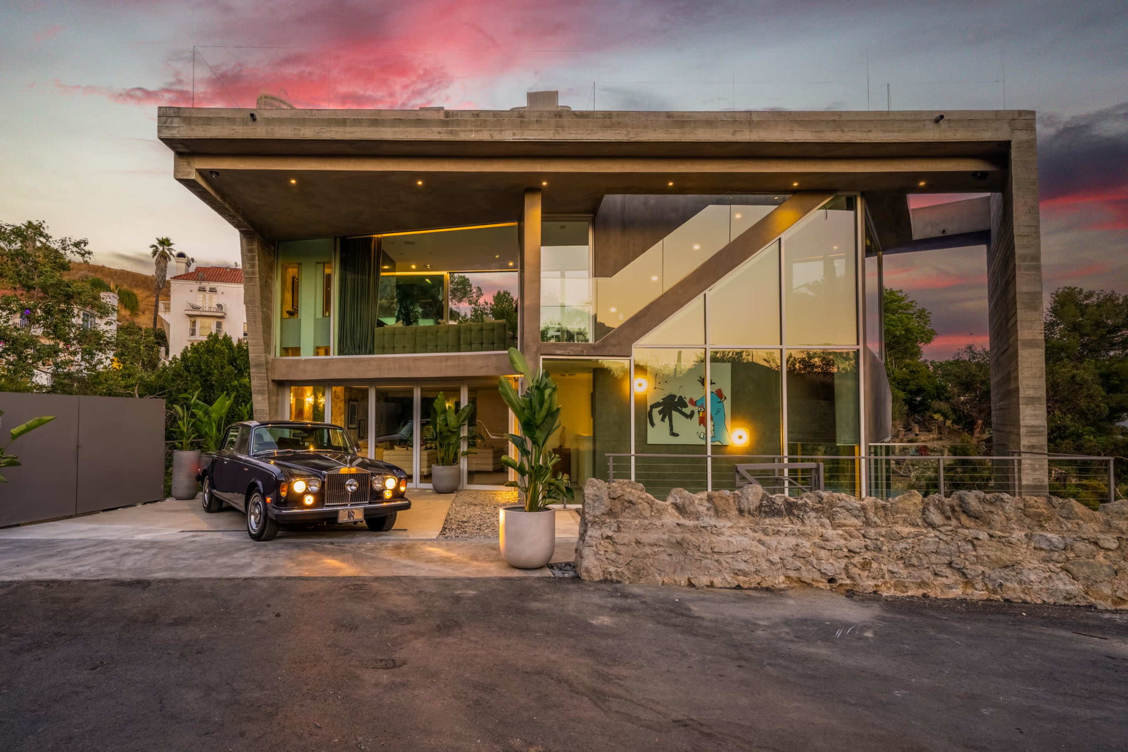 A modern house featuring large glass windows and a distinctive geometric design stands beside a vintage car parked on a gravel driveway, with a colorful sunset sky in the background.