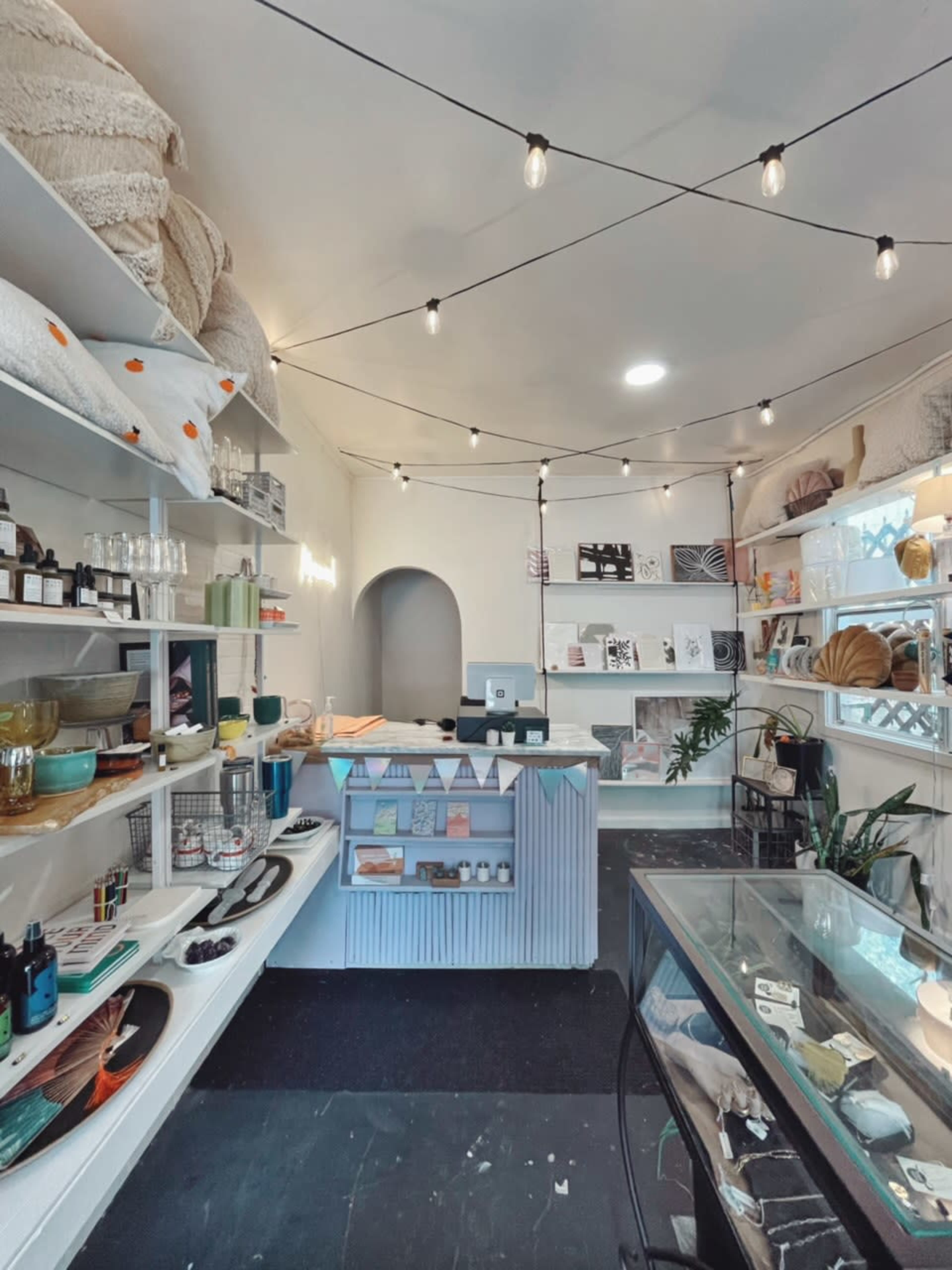 The image shows a neatly arranged retail space with shelves displaying various home goods and decor items, illuminated by string lights.