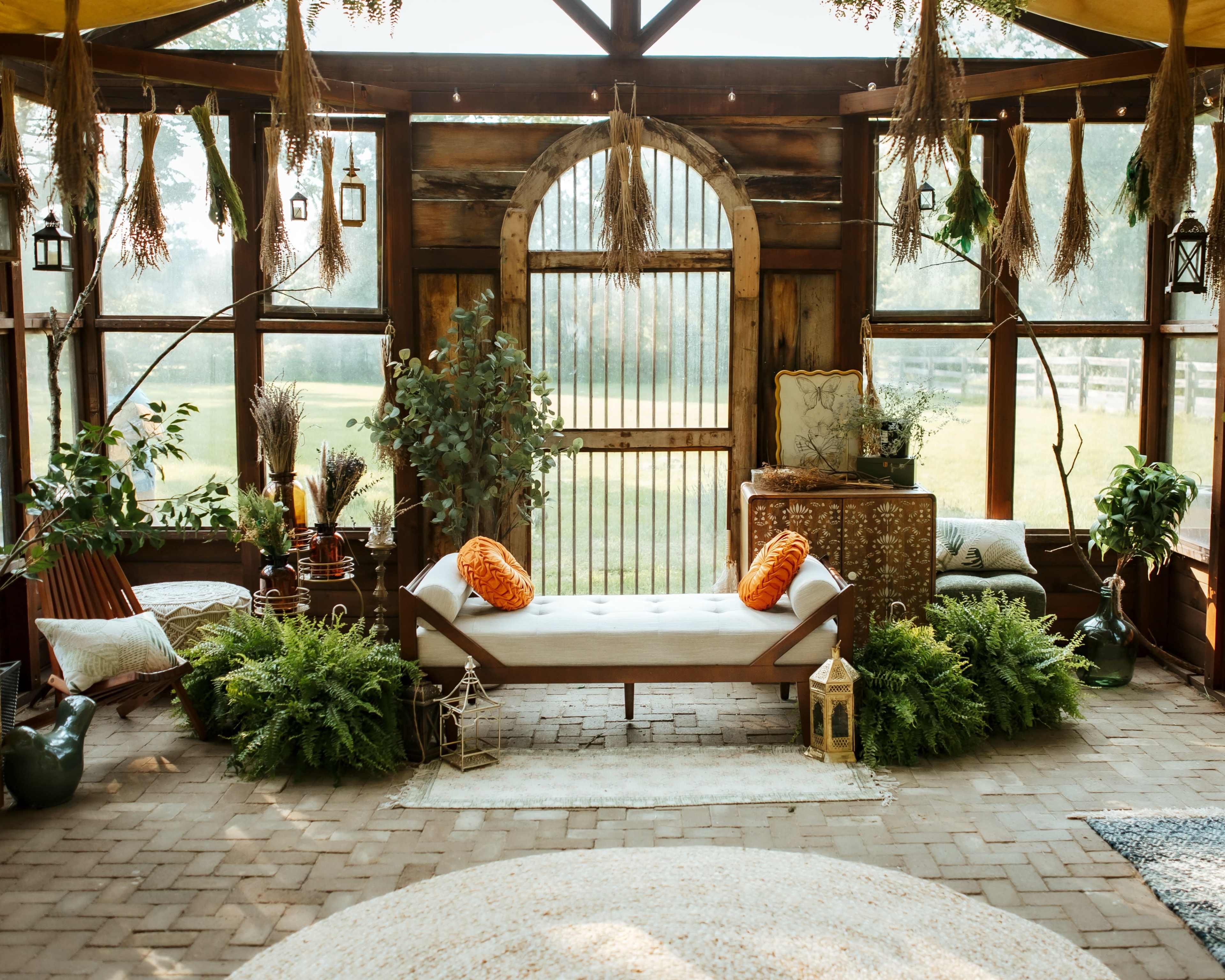 The image features a cozy indoor space with wooden walls, a daybed adorned with orange cushions, various plants, and decorative elements like lanterns and a gold-patterned table.