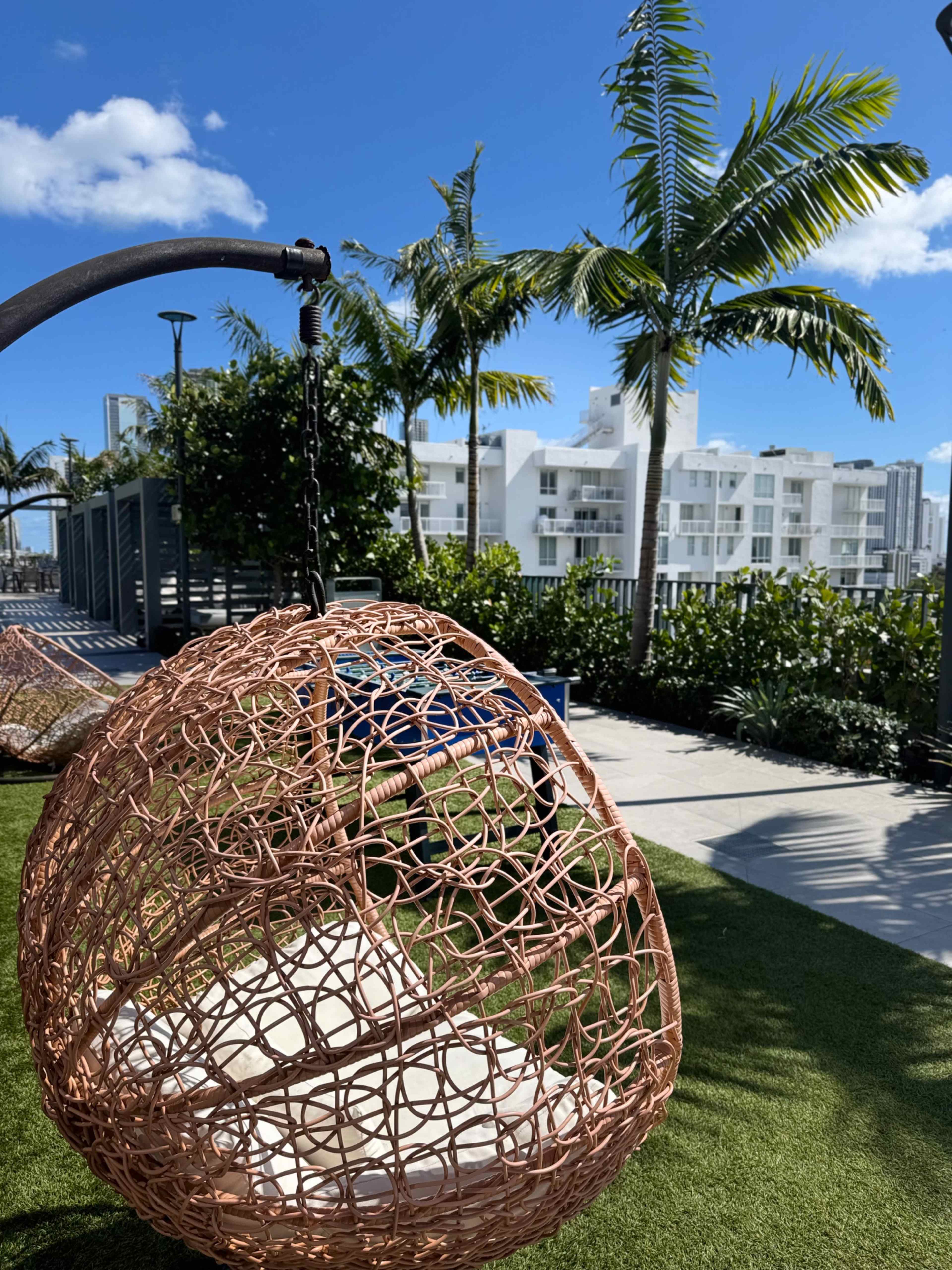 A wicker hanging chair is suspended in a sunny outdoor area surrounded by palm trees and modern buildings.