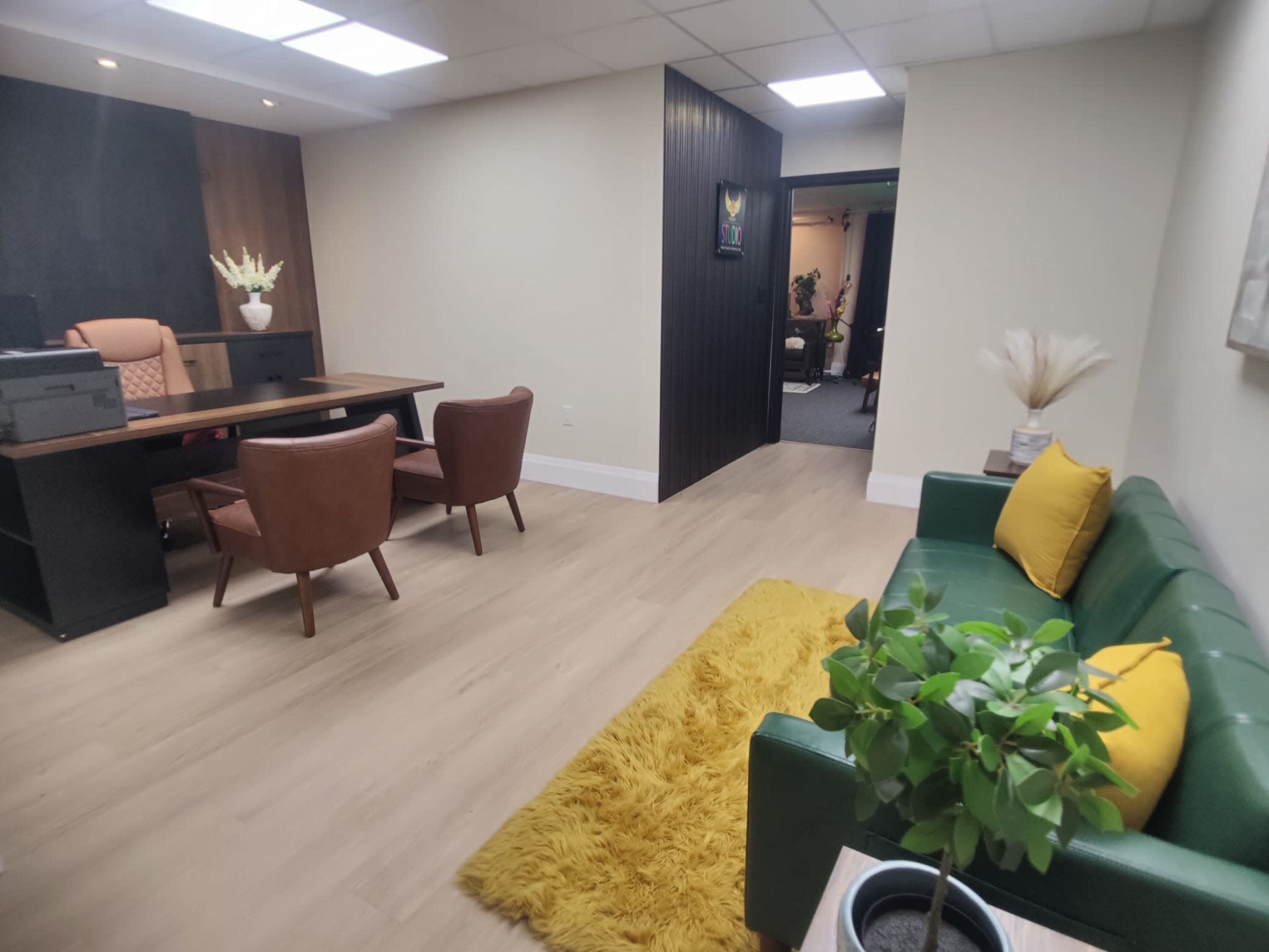 The image shows a modern office space featuring a desk with two chairs, a green couch with yellow cushions, a potted plant, and a rug on a wooden floor.