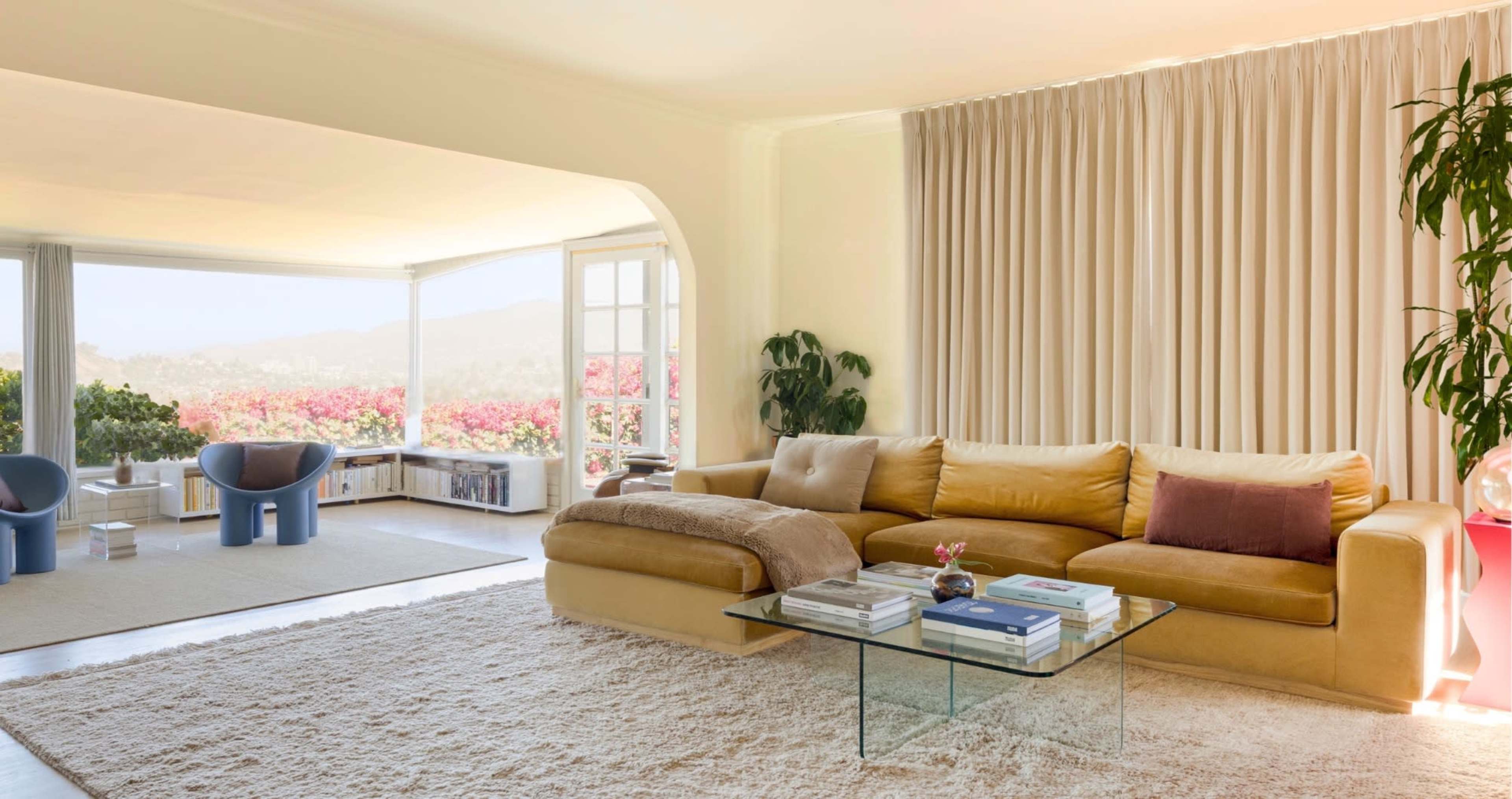 The image shows a modern living room with a yellow sectional sofa, a glass coffee table, and large windows featuring a view of a landscape filled with pink flowers.