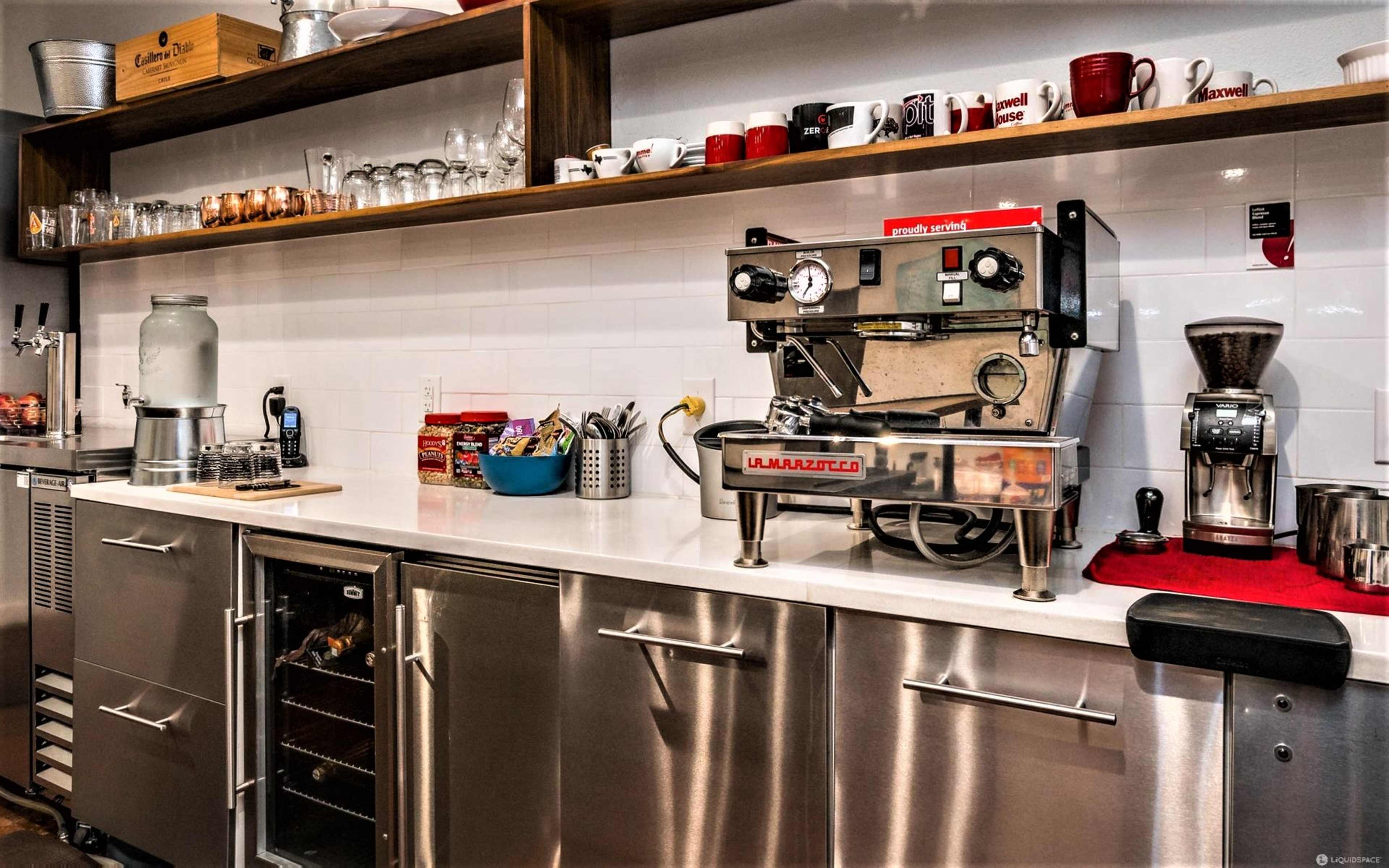 The image shows a modern coffee preparation area featuring an espresso machine, coffee grinder, and a collection of mugs on shelves above a stainless steel counter.