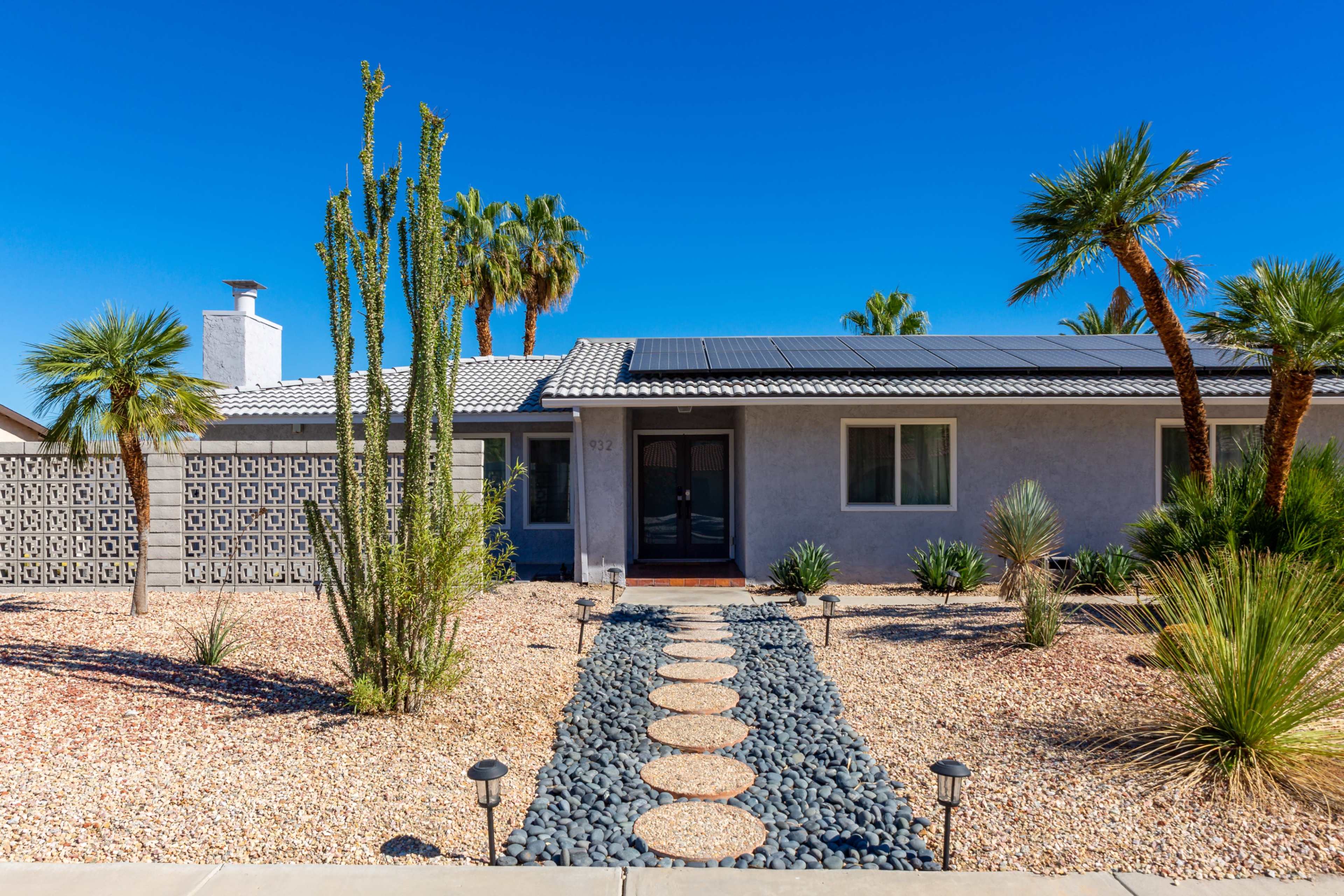 A modern single-story house with a landscaped front yard featuring various plants, a pathway made of stones, and solar panels on the roof.