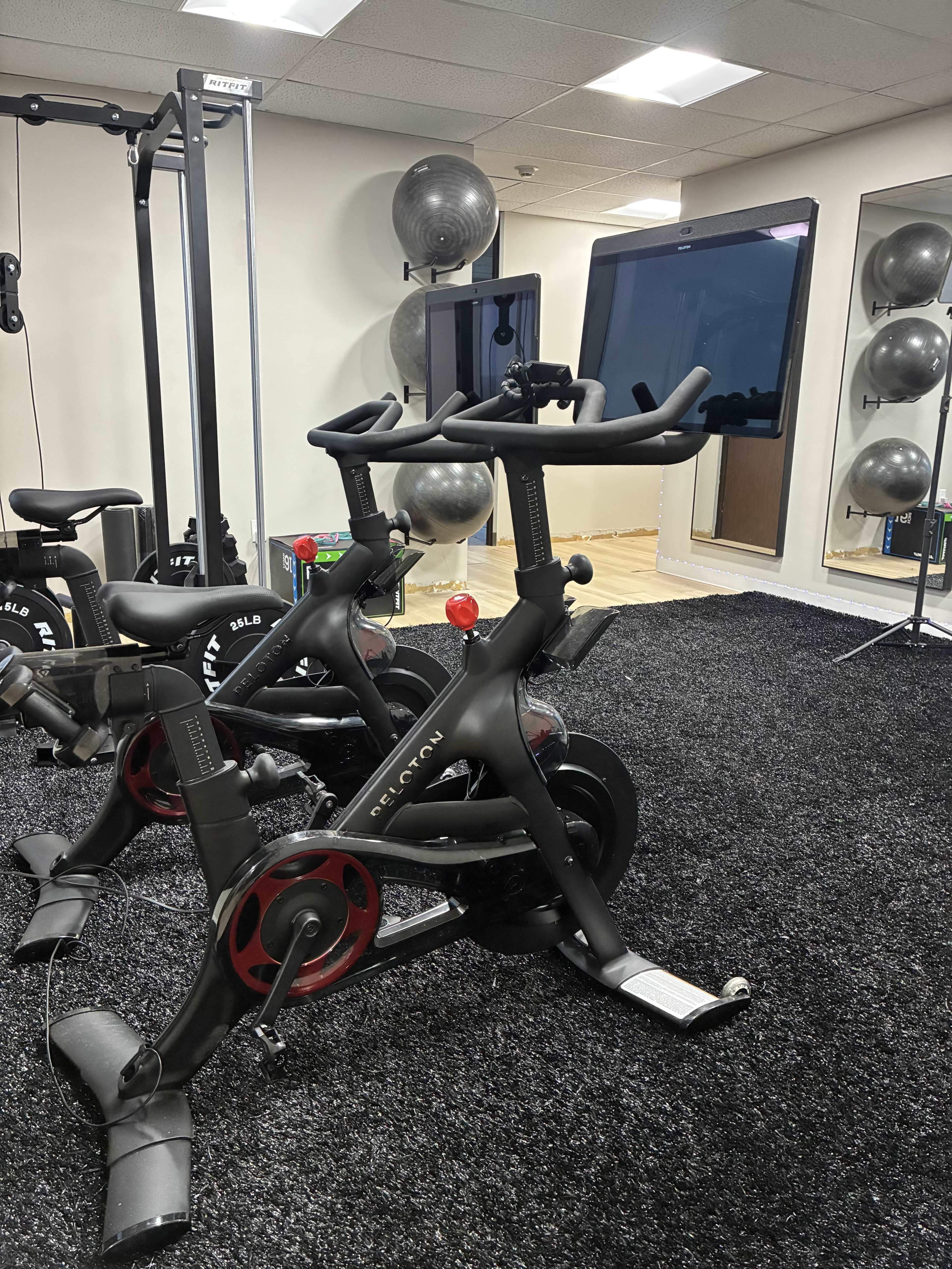 The image shows a fitness area with exercise bikes on a black carpeted floor, large mirrors, and exercise balls in the background.