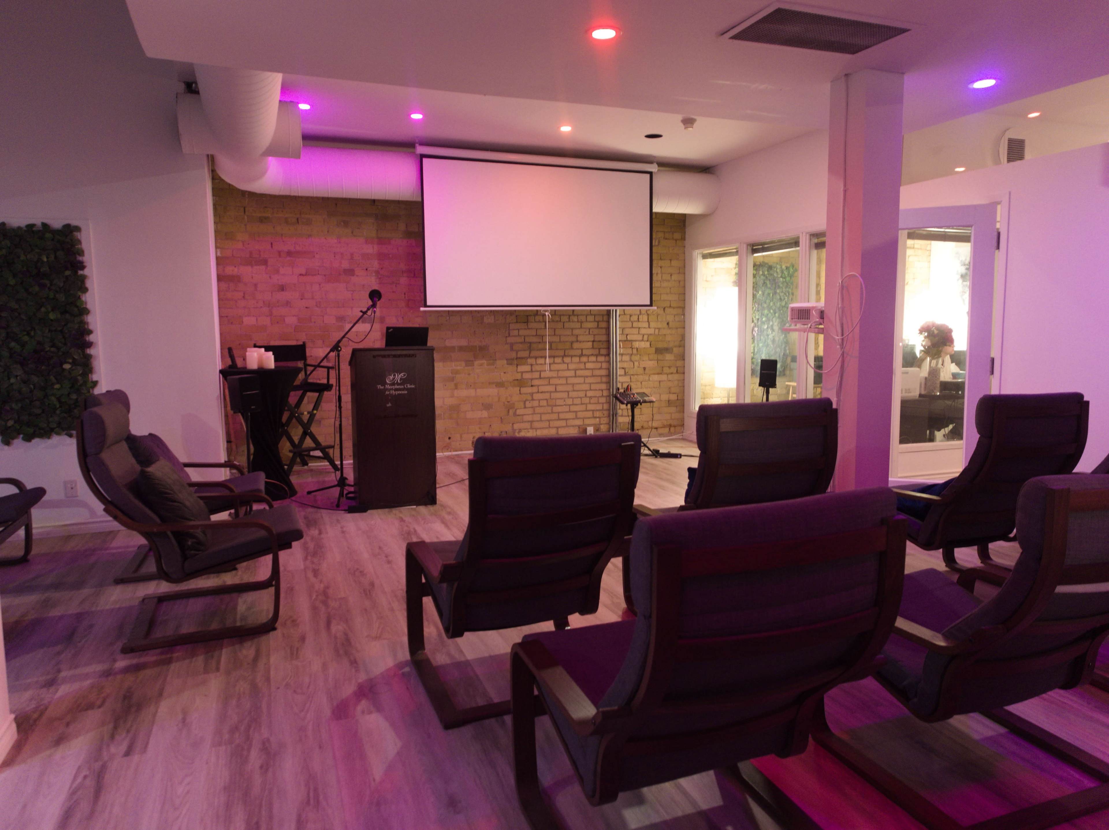 A small meeting room features several reclining chairs arranged facing a podium and a projector screen, with brick walls and ambient lighting.