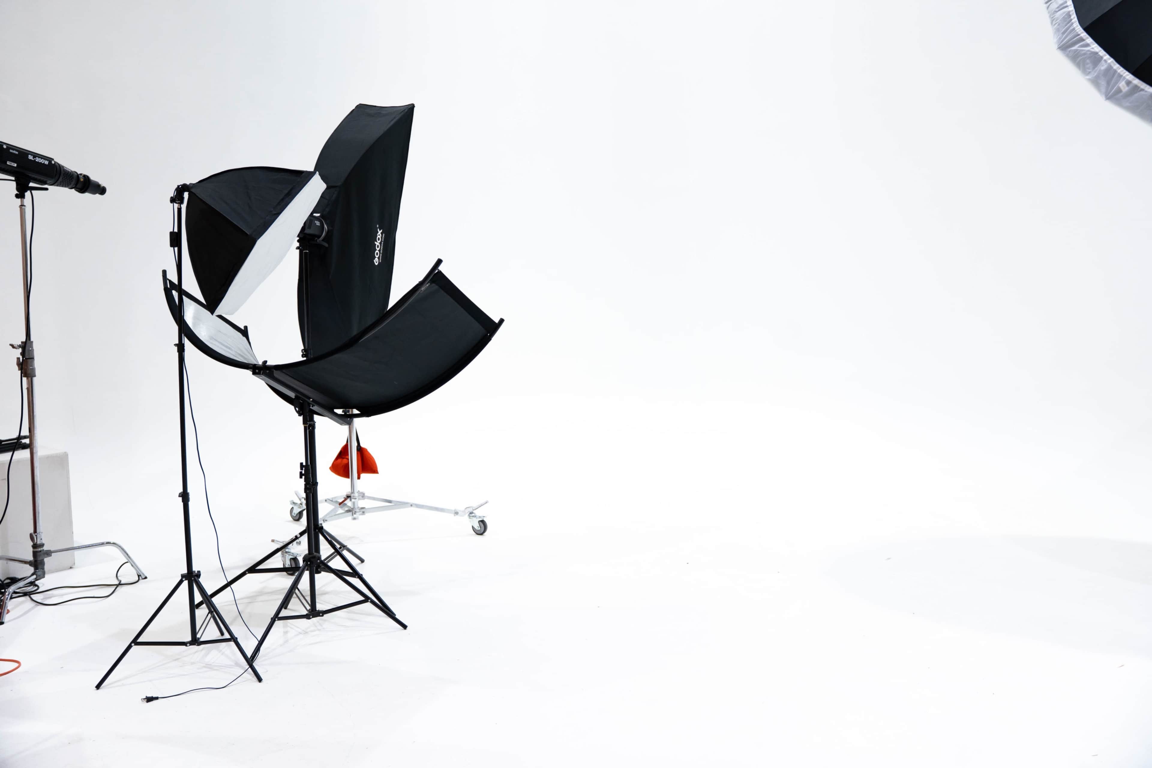 The image shows a photography studio setup with a black umbrella softbox, light stand, and a plain white backdrop.