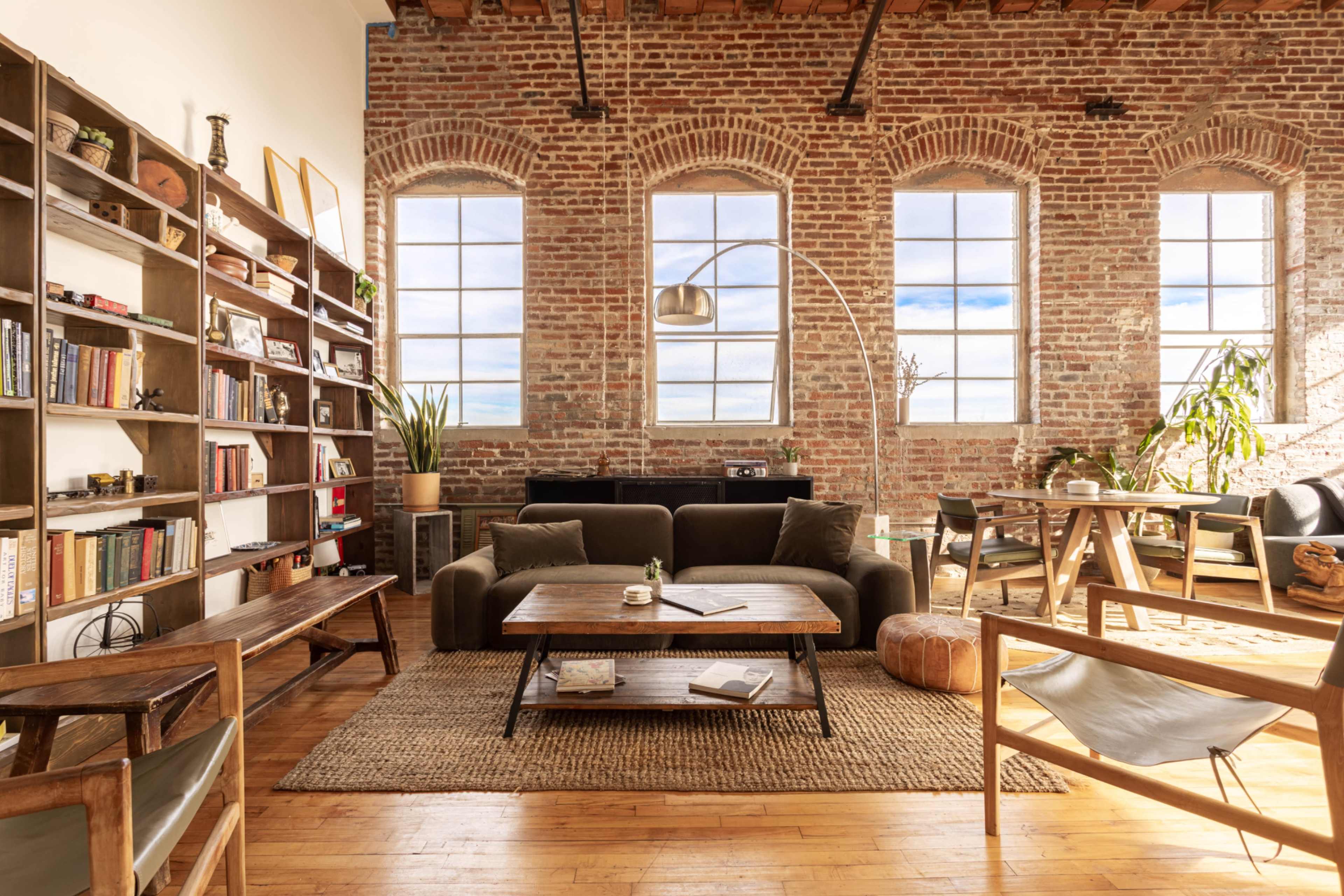 A cozy, industrial-style living space featuring exposed brick walls, large windows, a comfortable couch, a wooden coffee table, and various plants.
