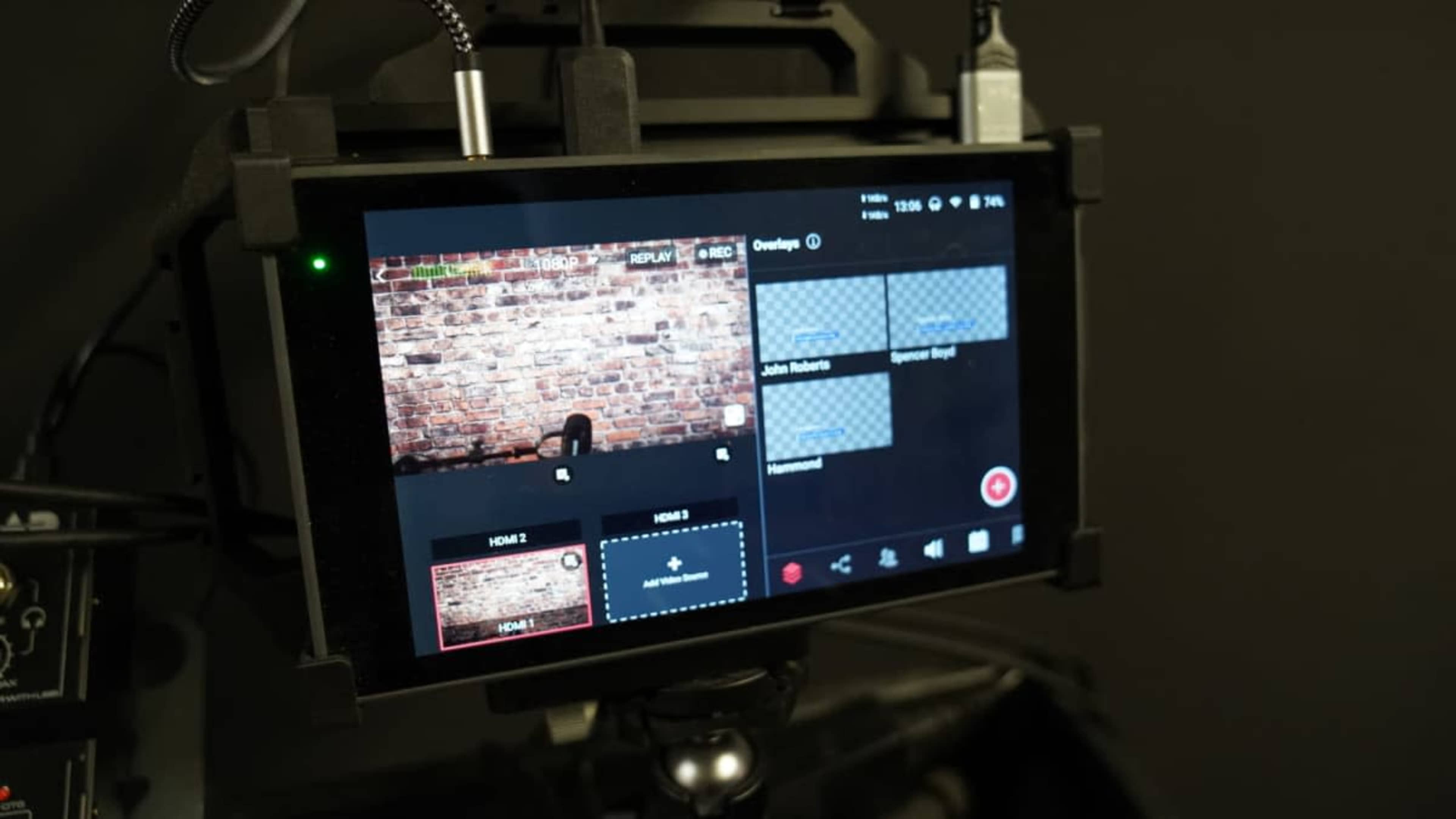 A digital monitor displays multiple video feeds, with a brick wall background visible on one screen and a camera setup below.