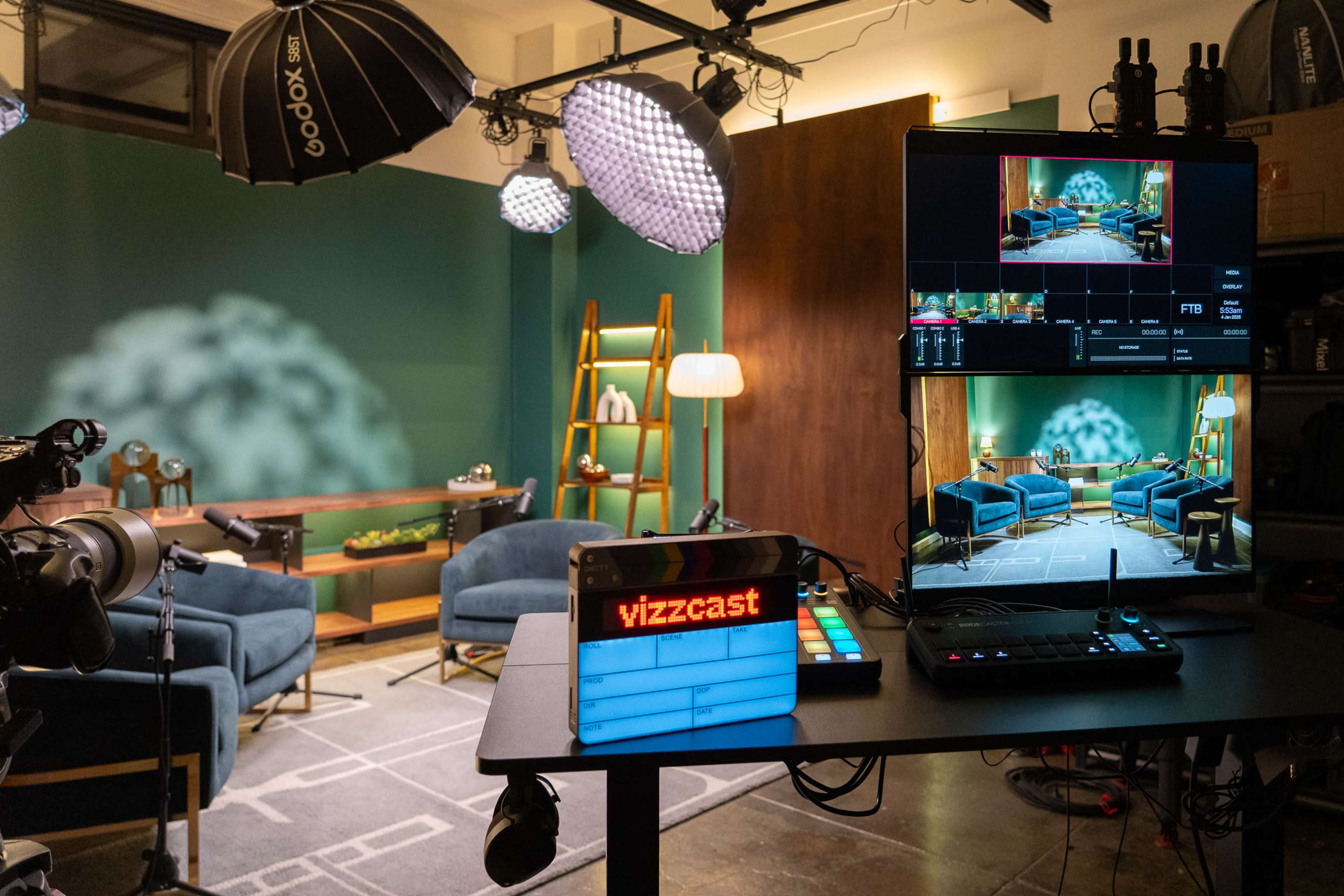 A modern studio setup features blue armchairs, a decorative backdrop, and a control monitor displaying a live feed.