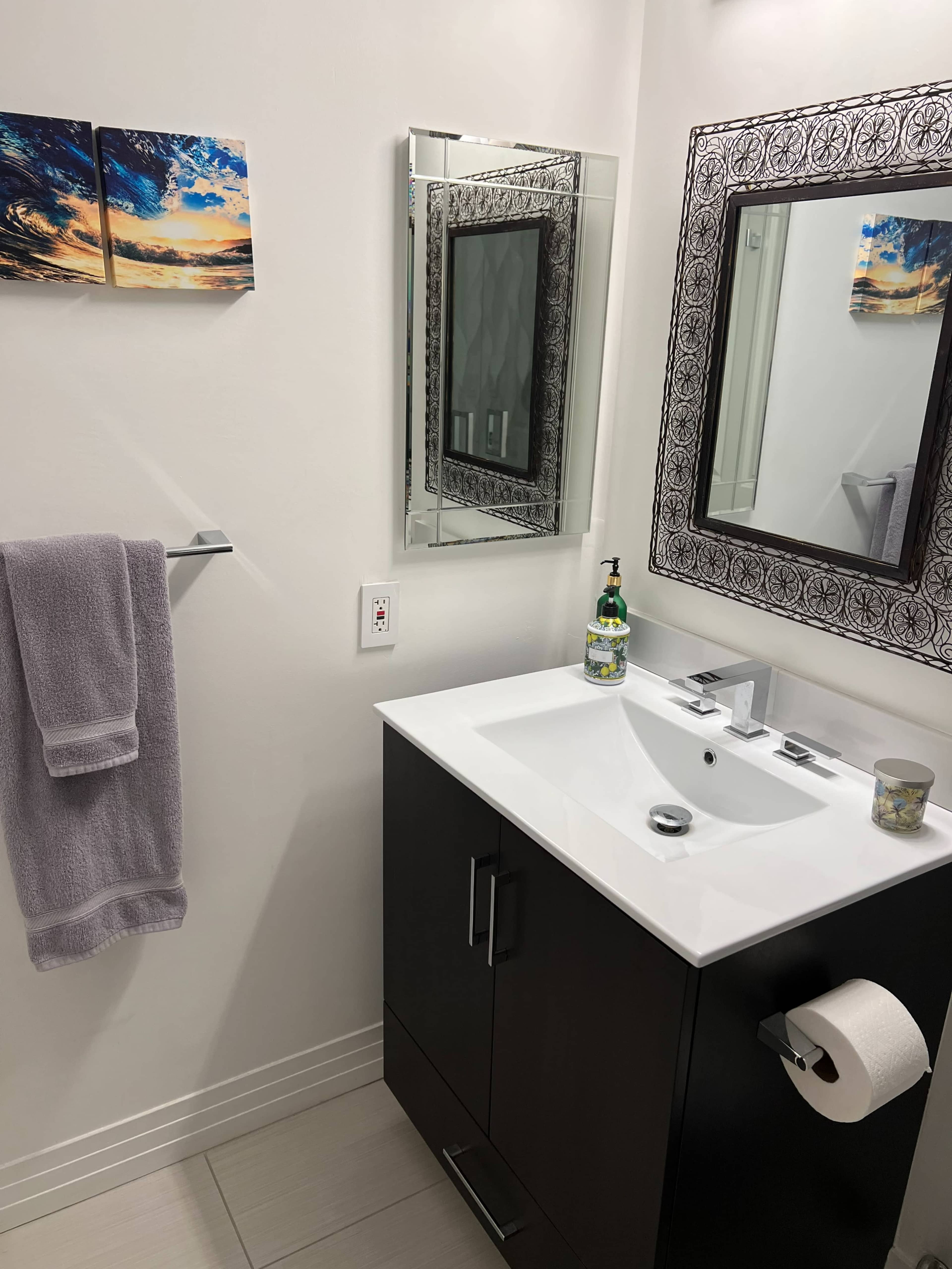 A modern bathroom with a sink and mirror, a towel hanging on the wall, and artwork above the sink.