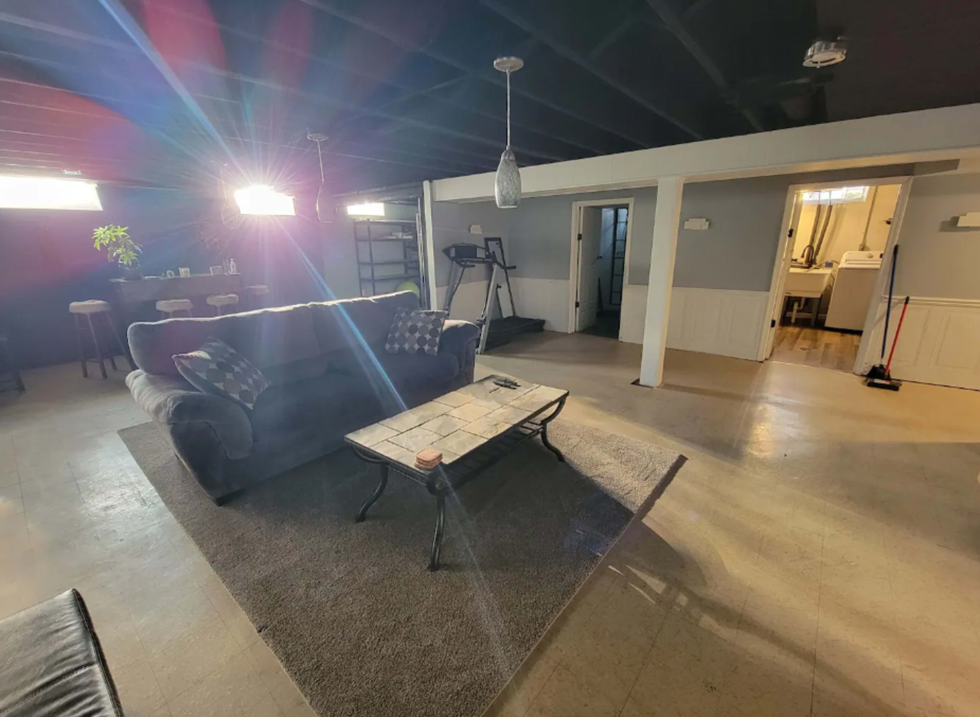 The image shows a spacious, well-lit basement living area with a couch, coffee table, and a view of a nearby utility space.