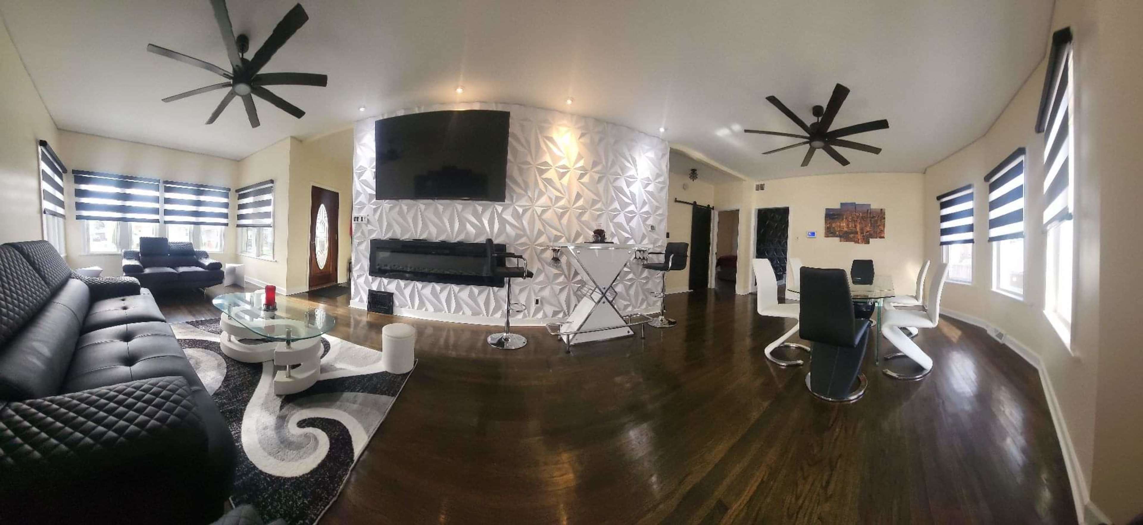 The image shows a modern, open-concept living and dining area furnished with black sofas, a glass table, and a metallic accent wall.