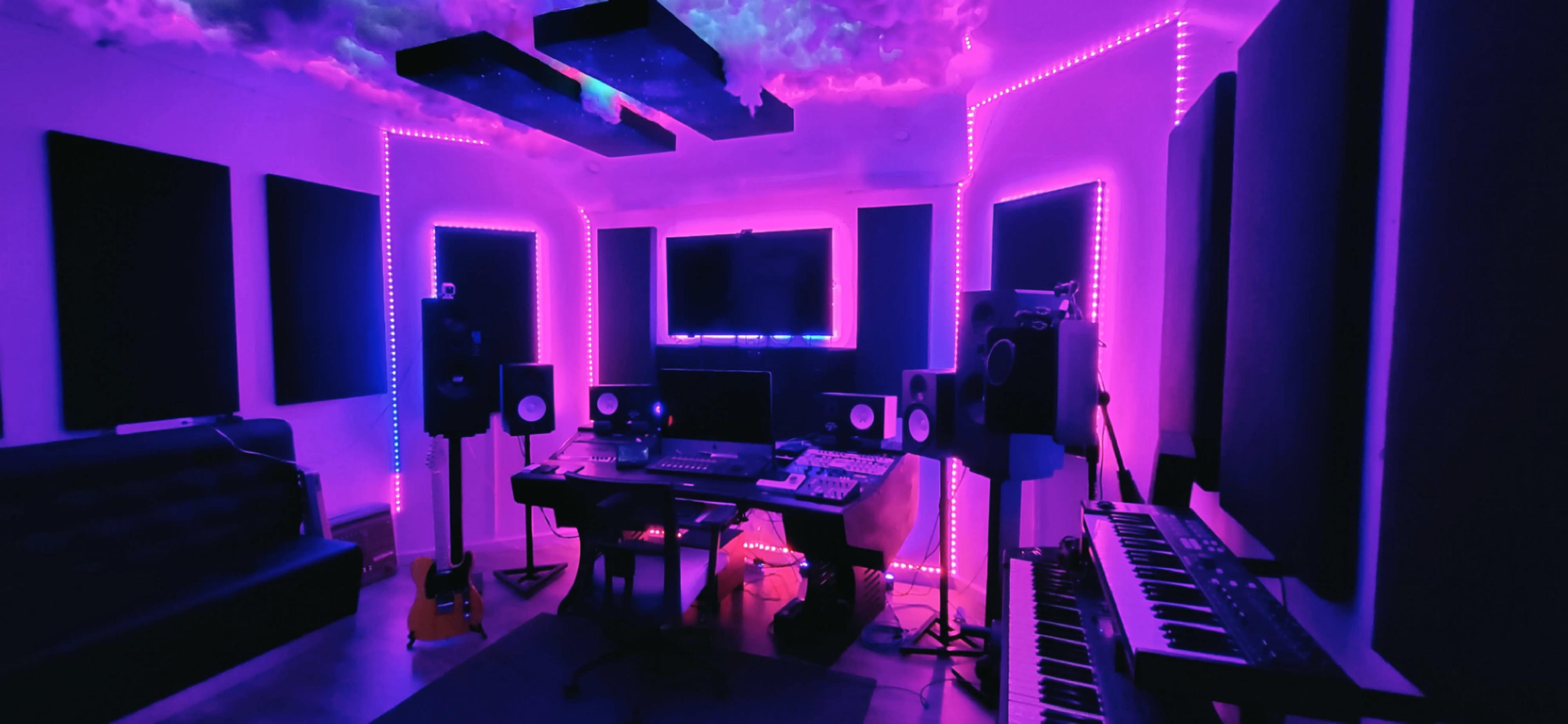 A music production studio featuring colorful LED lighting, a central desk with audio equipment, and soundproof panels on the walls.