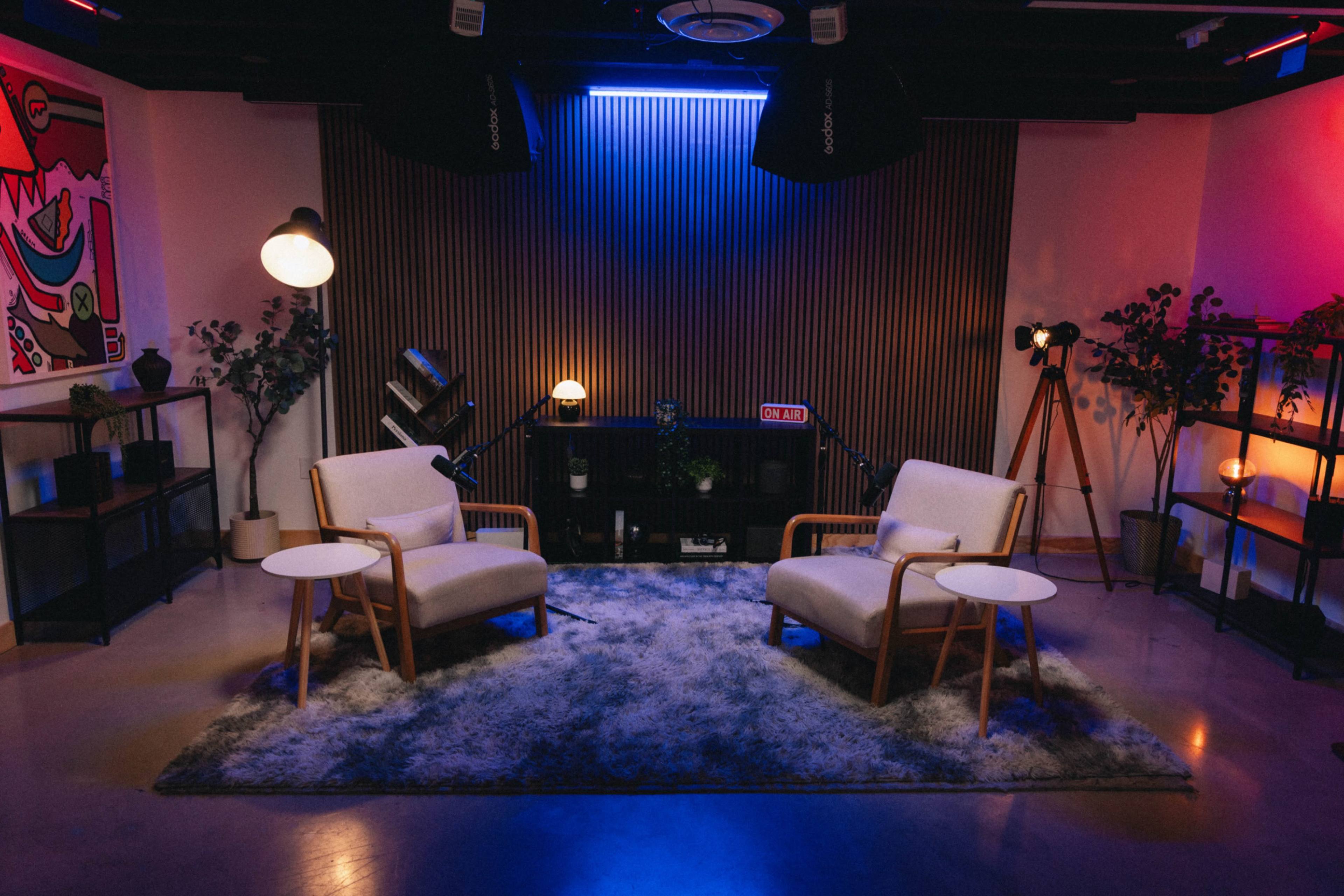 The image shows a modern interior with two light-colored armchairs, a small round table, and various plants, all arranged on a large rug under ambient lighting.
