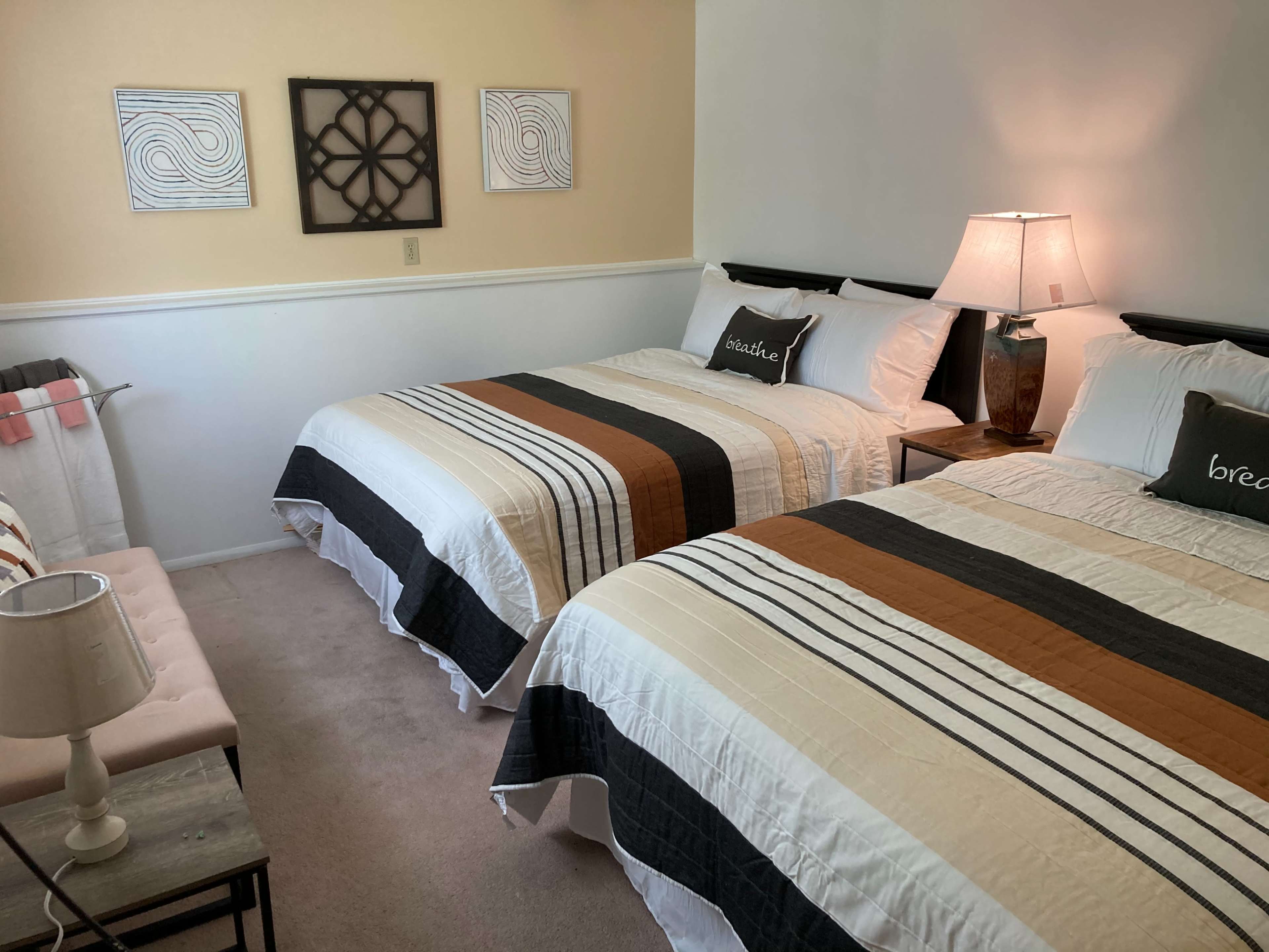 A room with two neatly made beds, each featuring striped bedding, a bedside lamp, and decorative wall art.