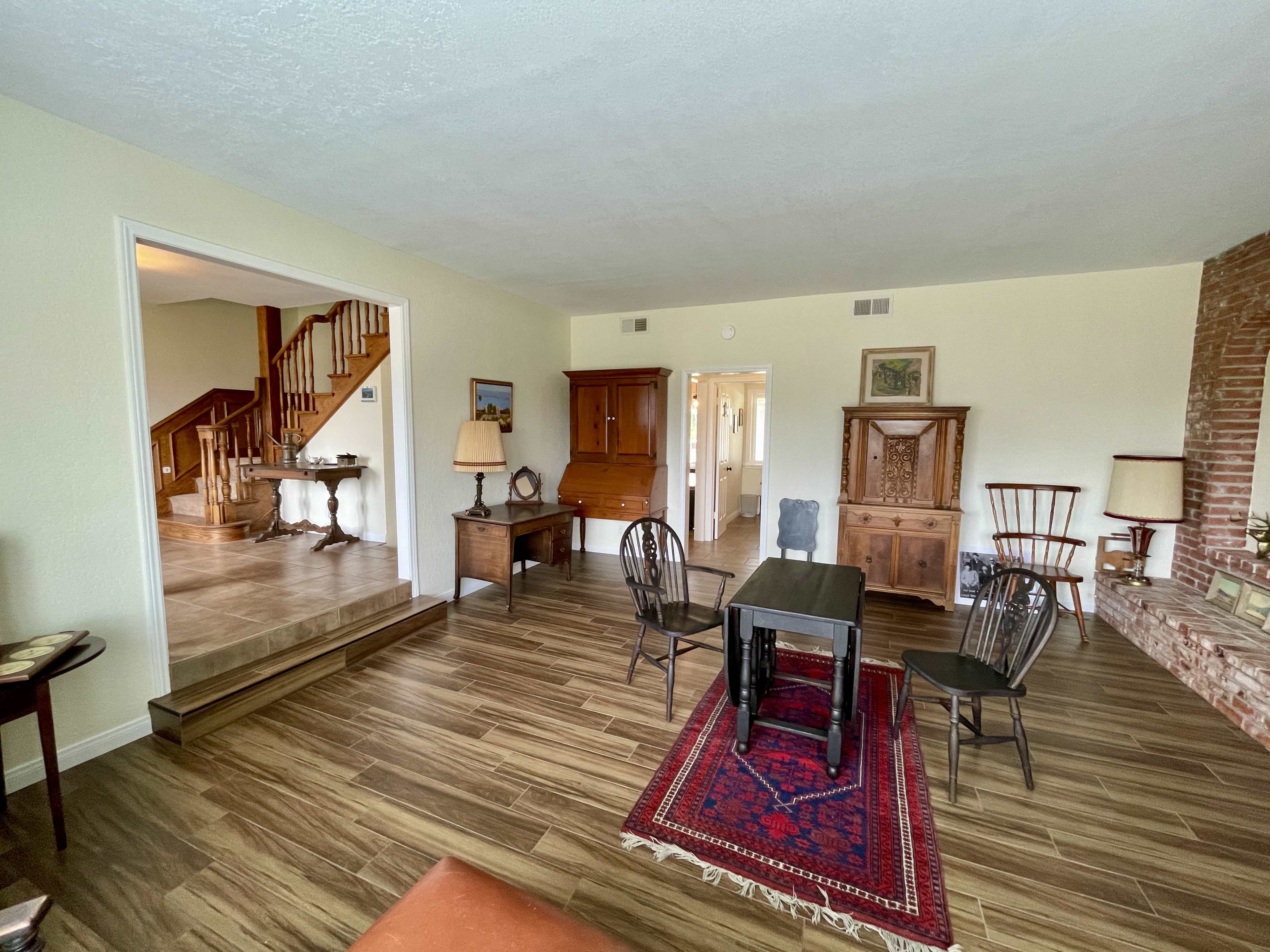The image shows a spacious living room featuring a table and chairs, a rug, and wooden furniture, with stairs visible in the background.