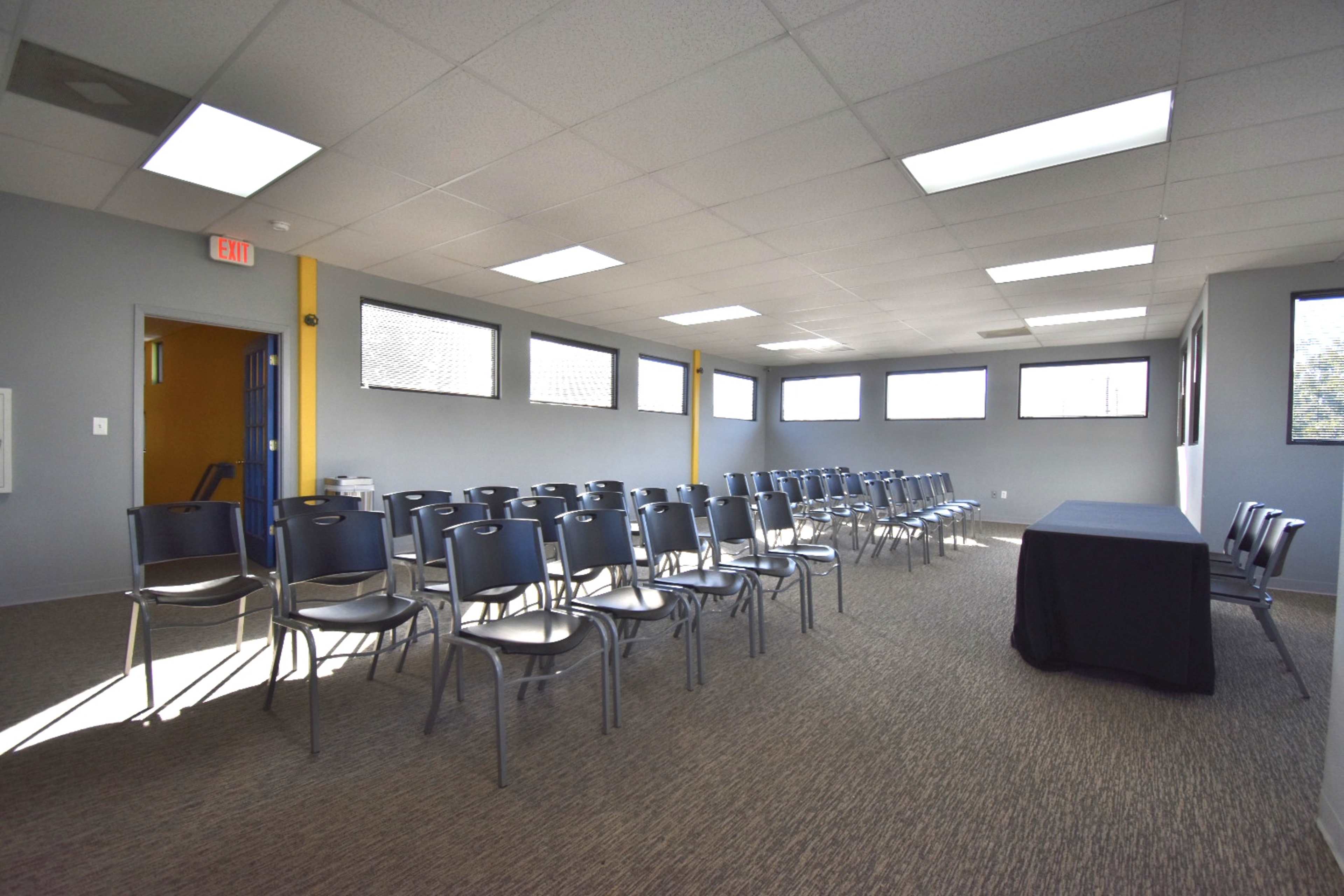 A row of chairs is arranged in a meeting room with a black table at the front and windows allowing natural light.