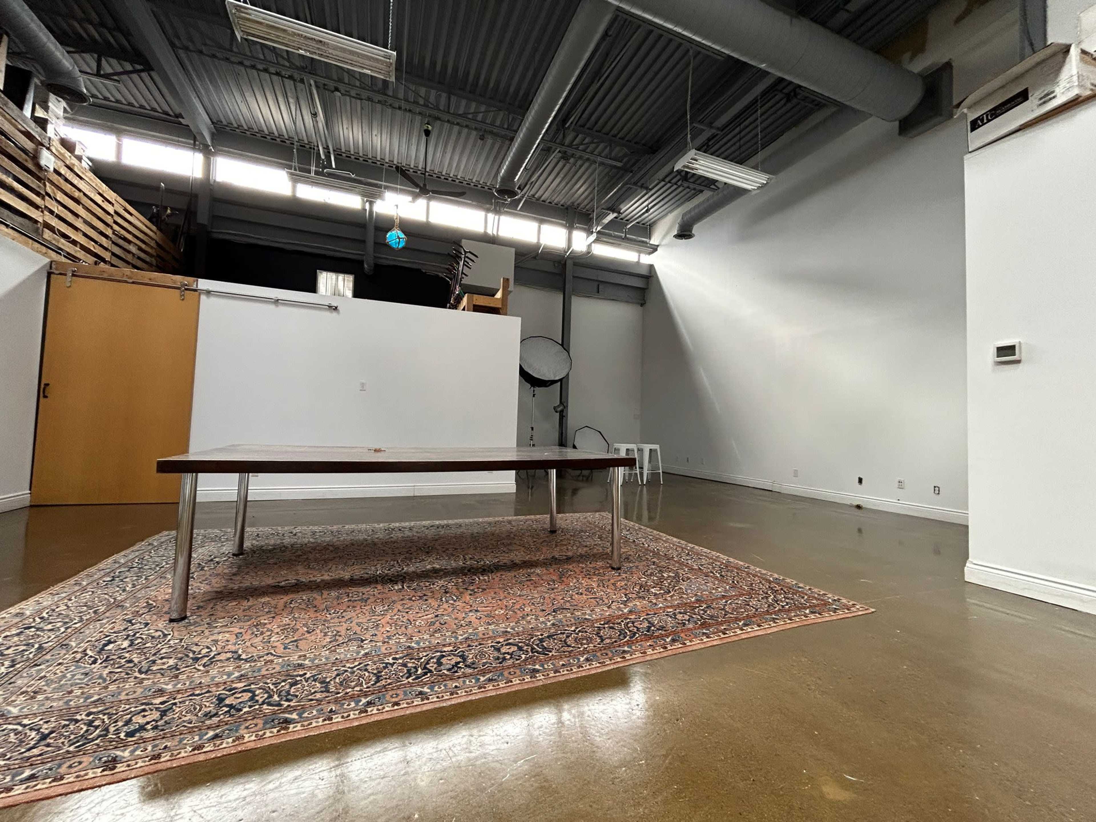 An empty room features a wooden table on a rug, with a backdrop visible in the distance.