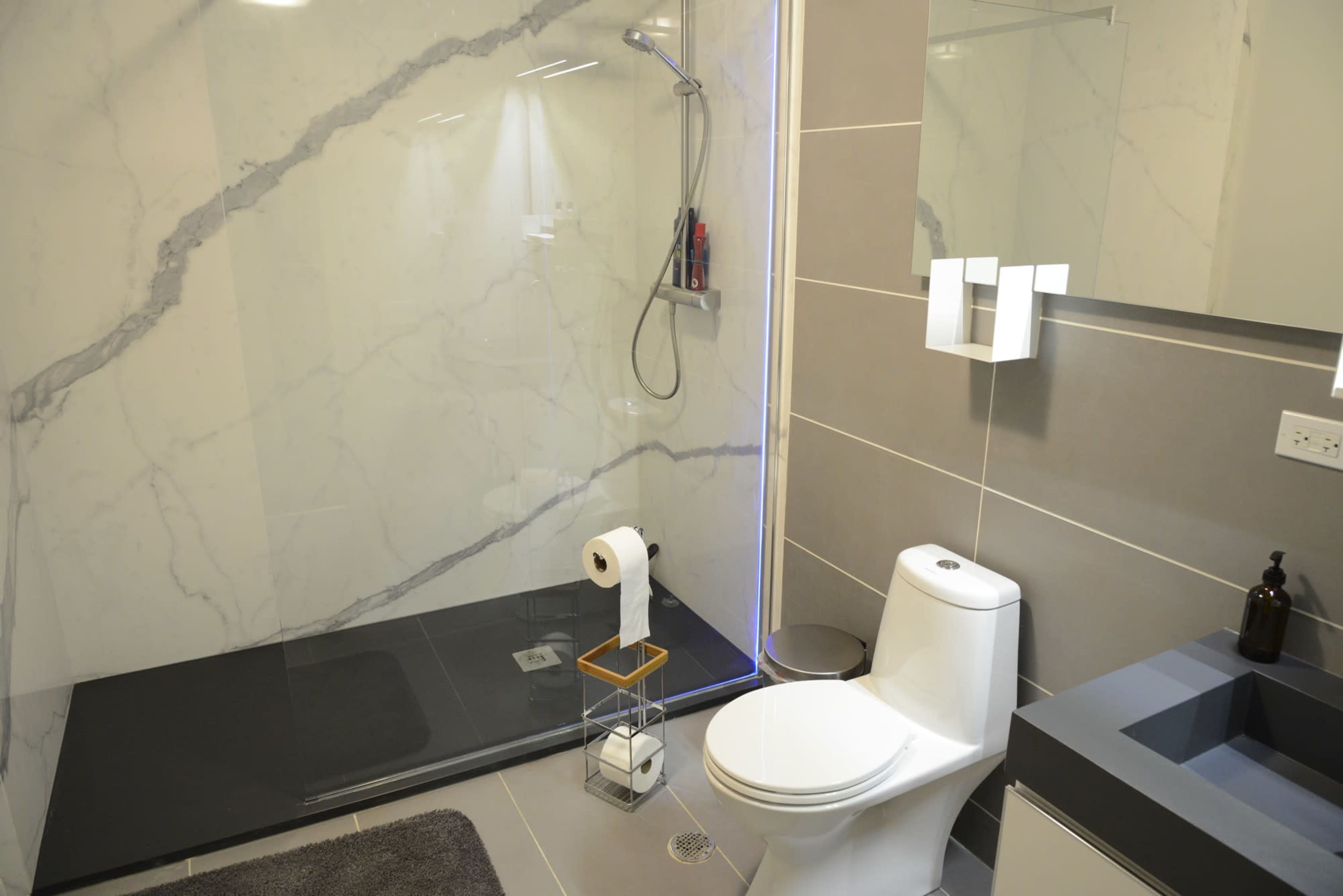 The image shows a modern bathroom featuring a glass shower enclosure, a toilet, a sink with a dark countertop, and a towel rack.