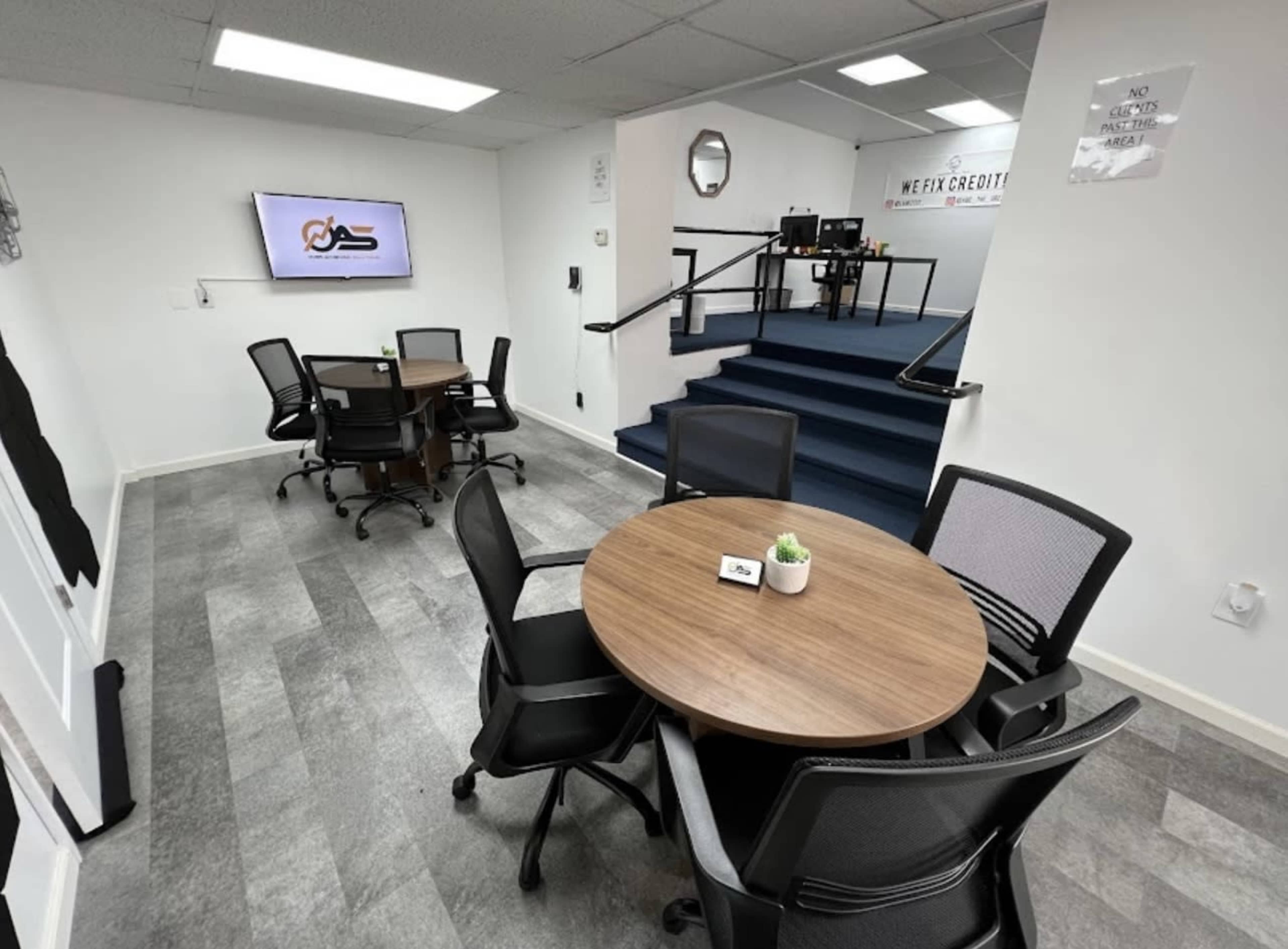 A modern office space with two round wooden tables and black mesh chairs, along with a staircase leading to a separate area.