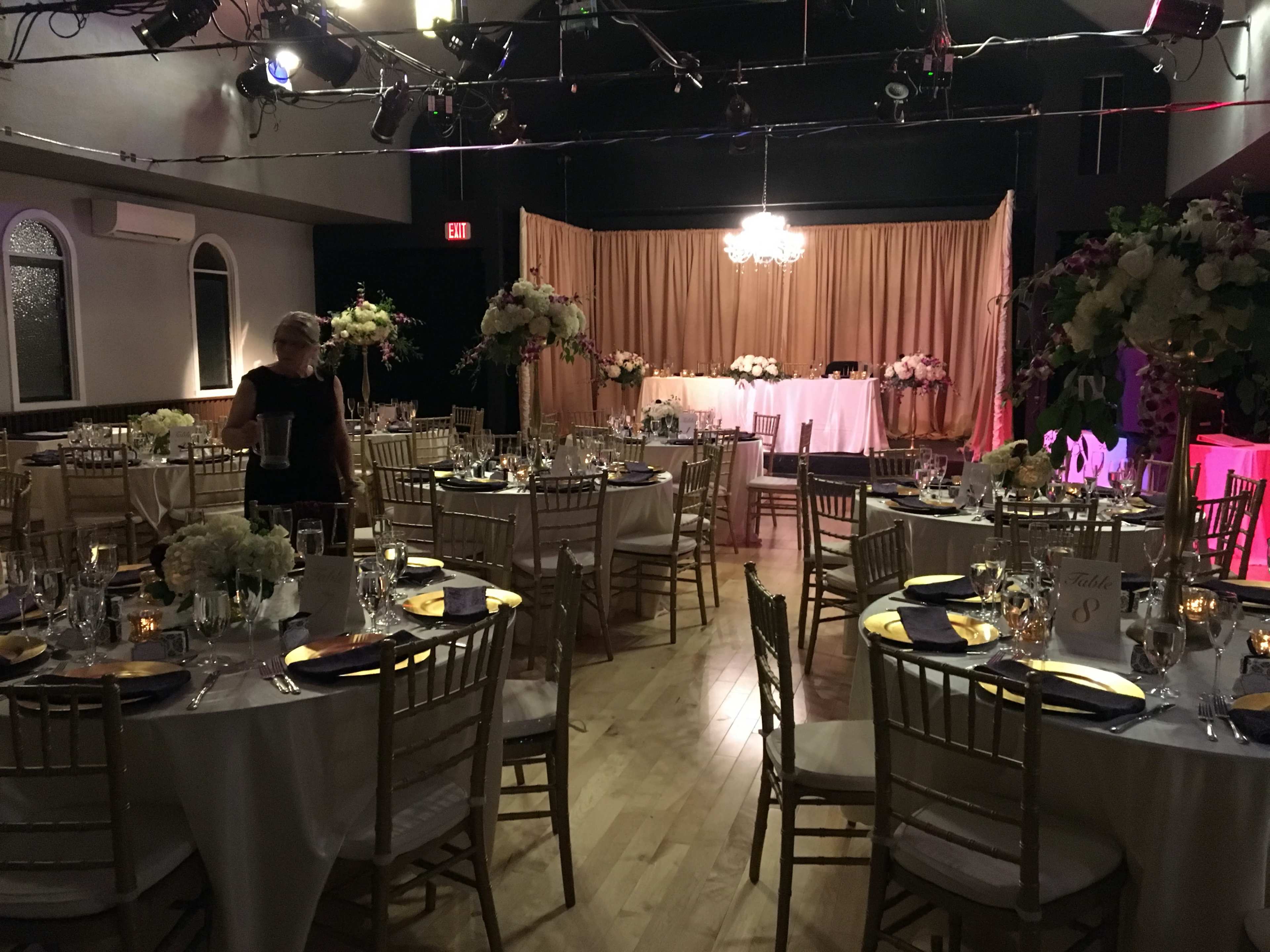 A banquet hall is set up for a formal event, featuring round tables with floral centerpieces and a stage adorned with a draped backdrop.