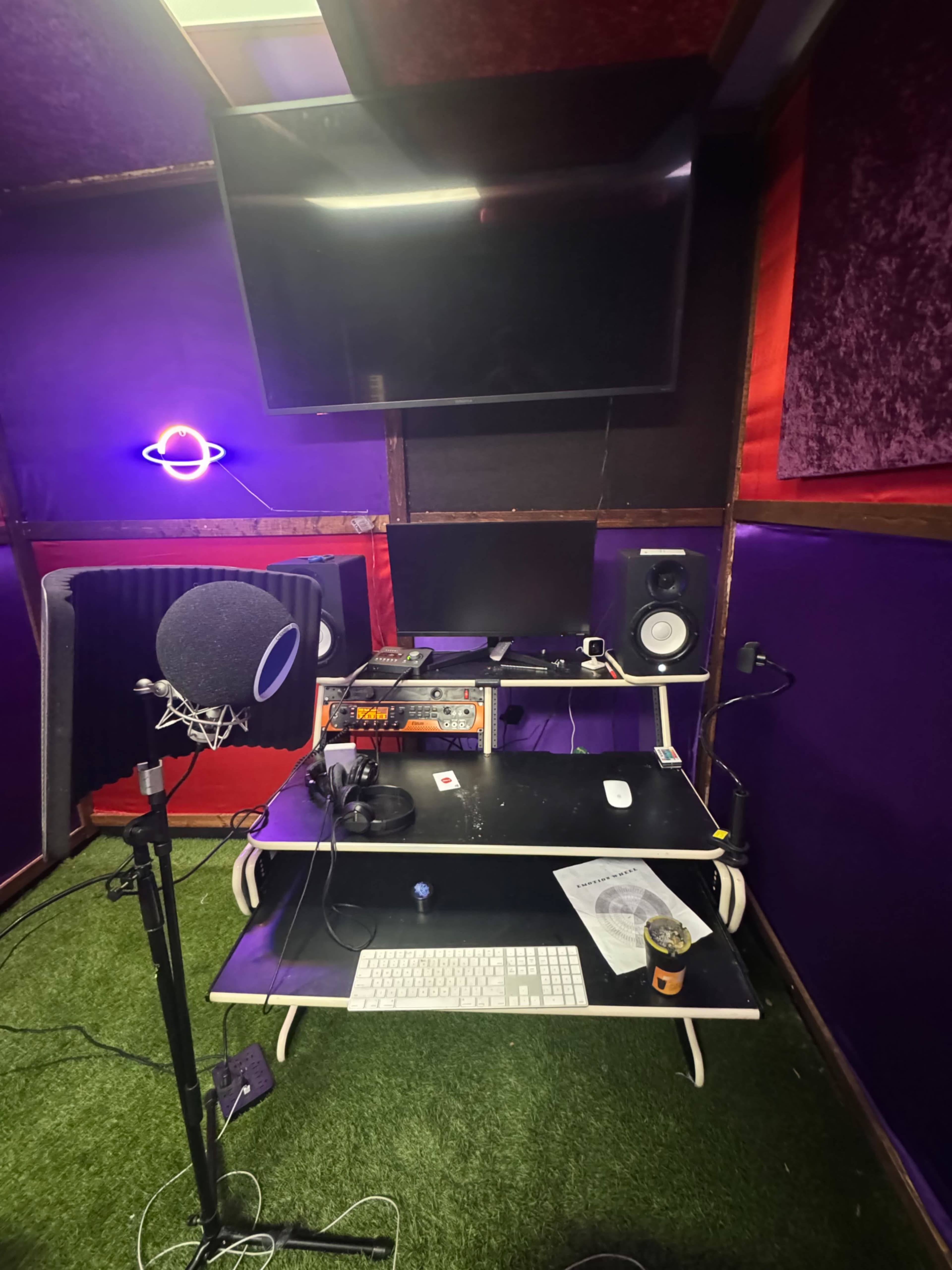 The image shows a small music recording studio with a microphone on a stand, a computer desk equipped with audio monitors, and a large screen on the wall.