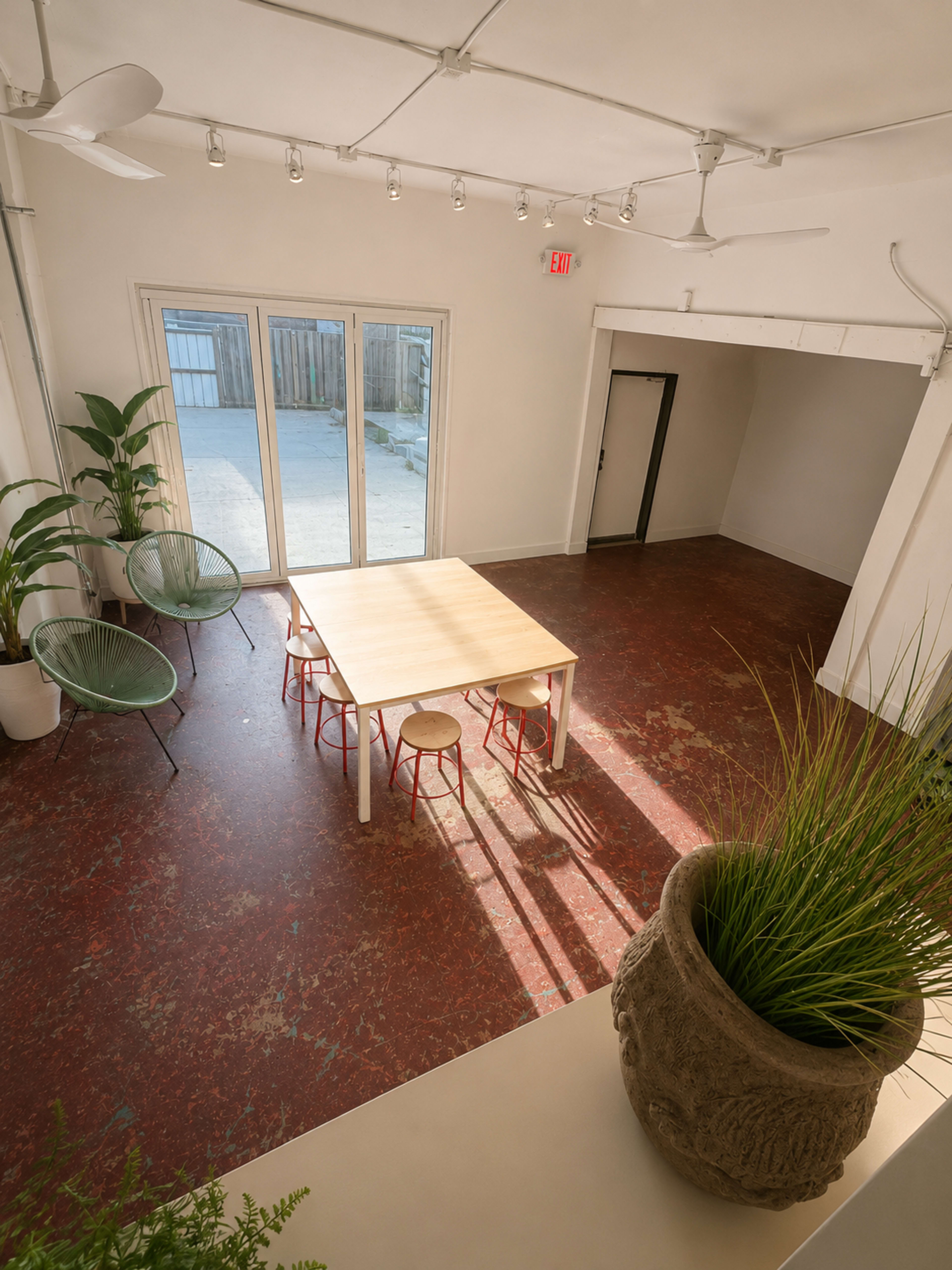 The image shows a spacious, brightly lit room with a wooden table surrounded by four chairs, potted plants, and large glass doors leading to an outdoor area.