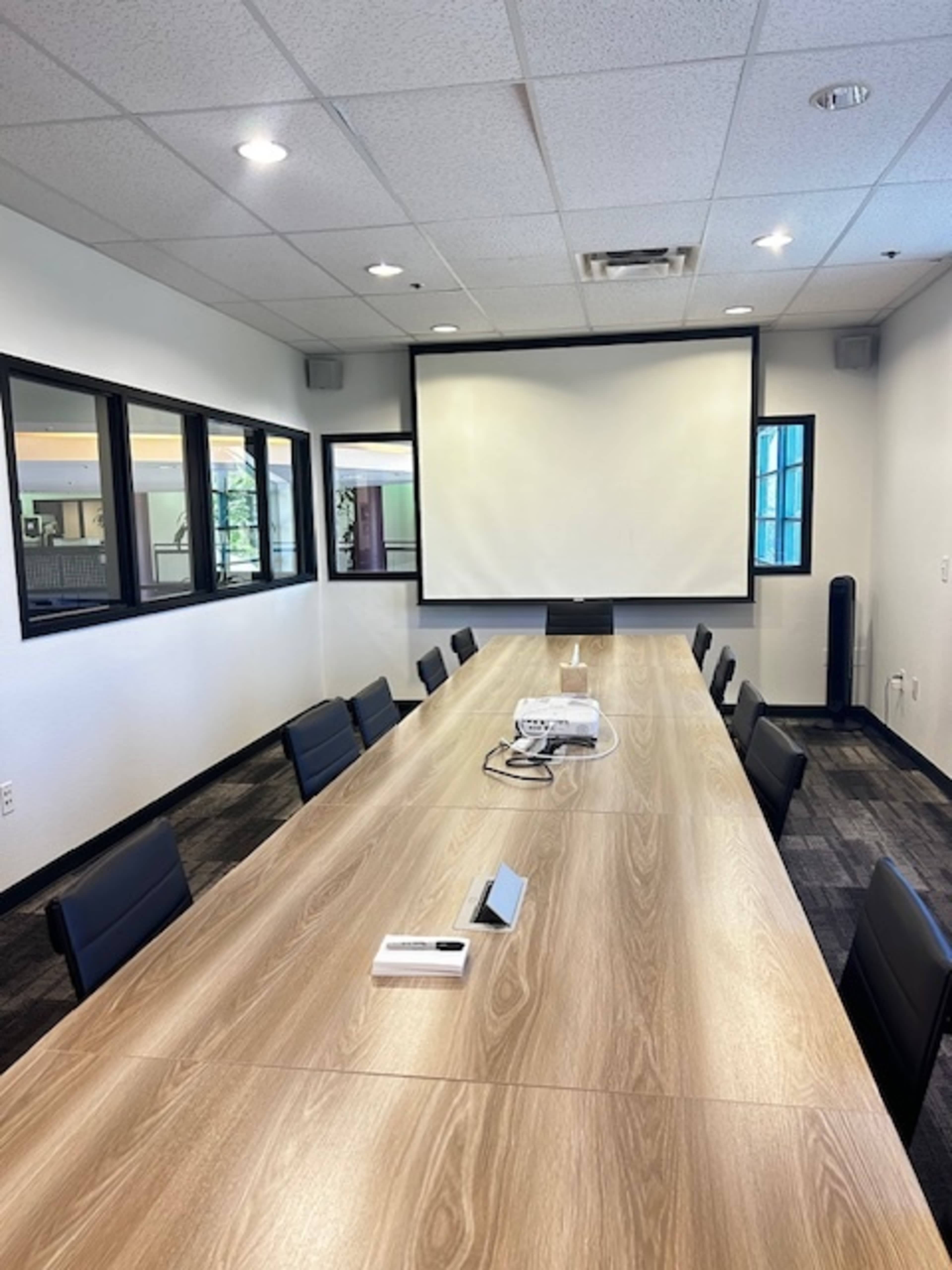 A large conference room featuring a long table surrounded by black chairs, with a projection screen at one end and windows along the walls.