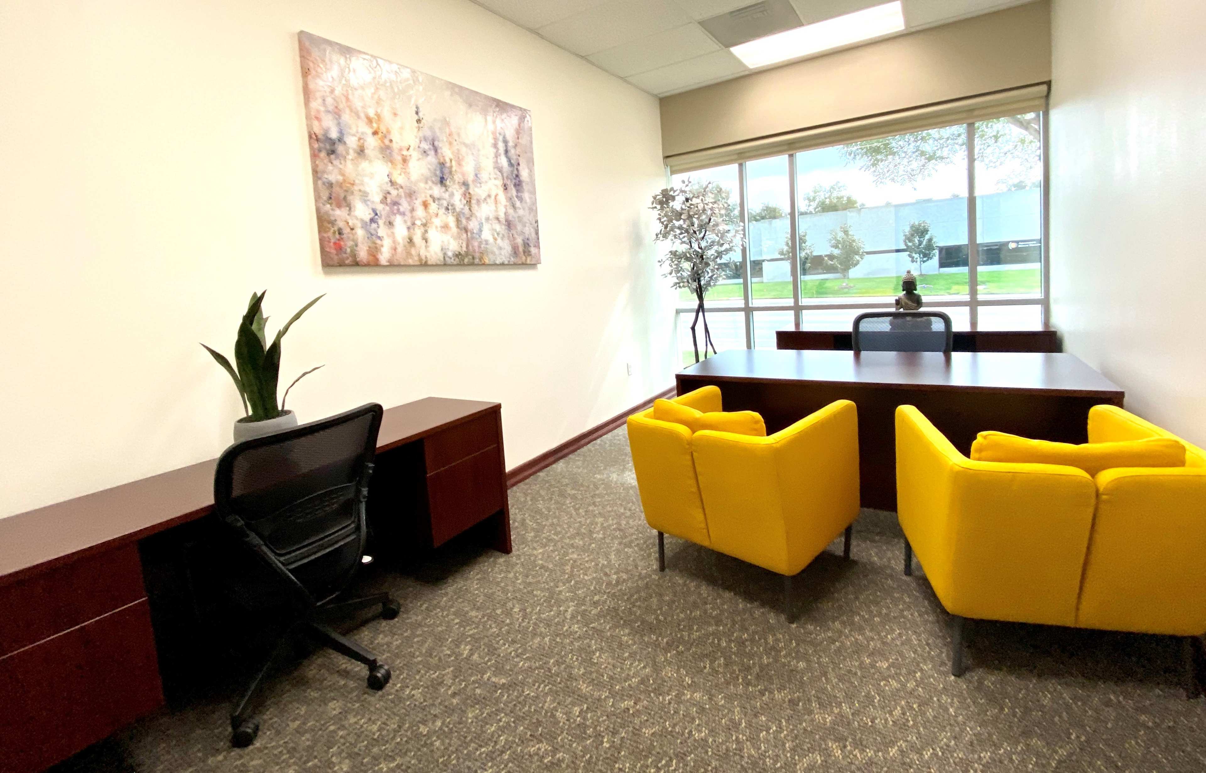The image shows an office space featuring a large window with greenery outside, a large desk with two bright yellow chairs, and a potted plant on the side.