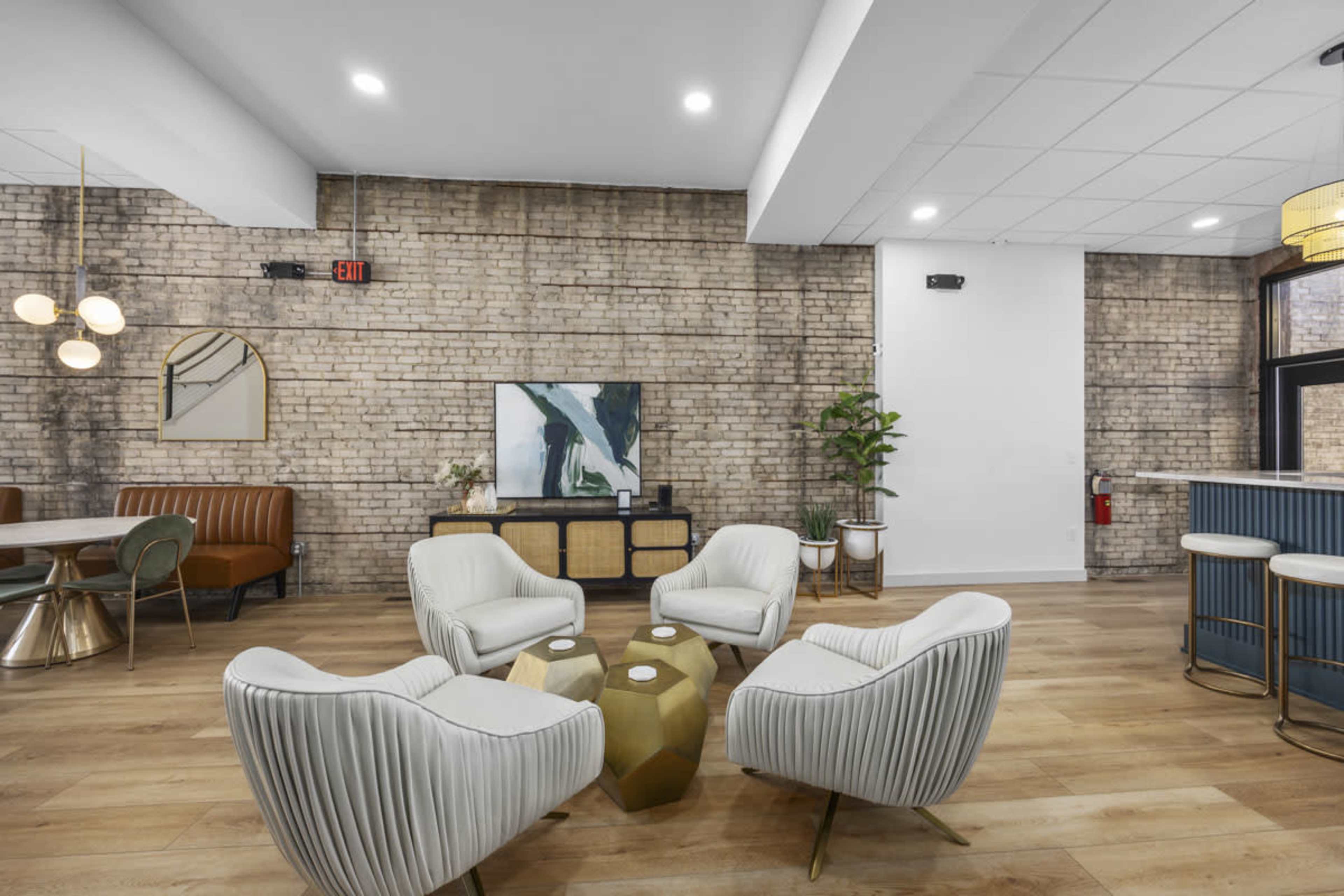 The image shows a modern lounge area with four white chairs arranged around a hexagonal coffee table, set against a backdrop of exposed brick walls and wooden flooring.