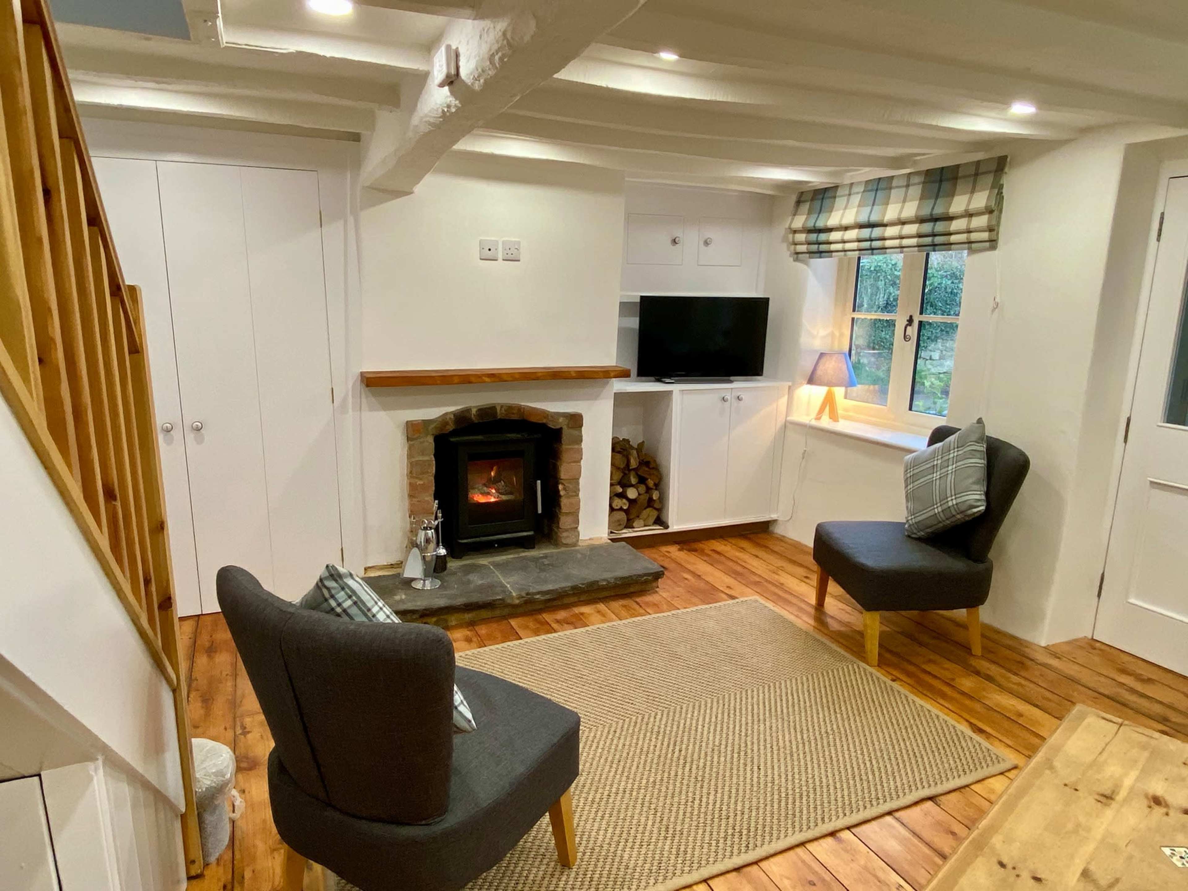A cozy living room with two chairs facing a stone fireplace, a television mounted on the wall, and a window dressed in plaid curtains.