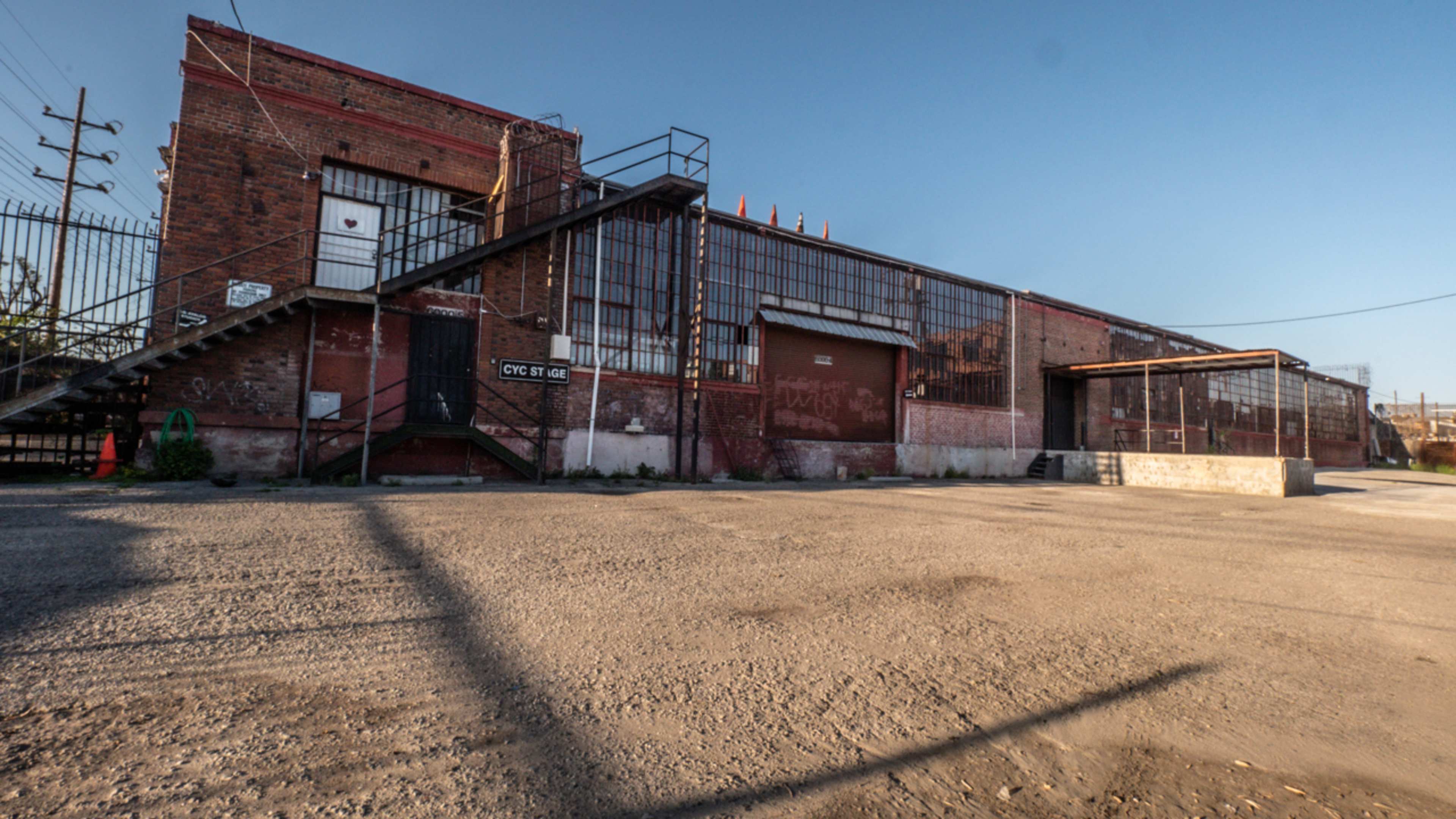 The image shows a large, red-brick industrial building with a metal staircase and several windows, set against a clear blue sky.