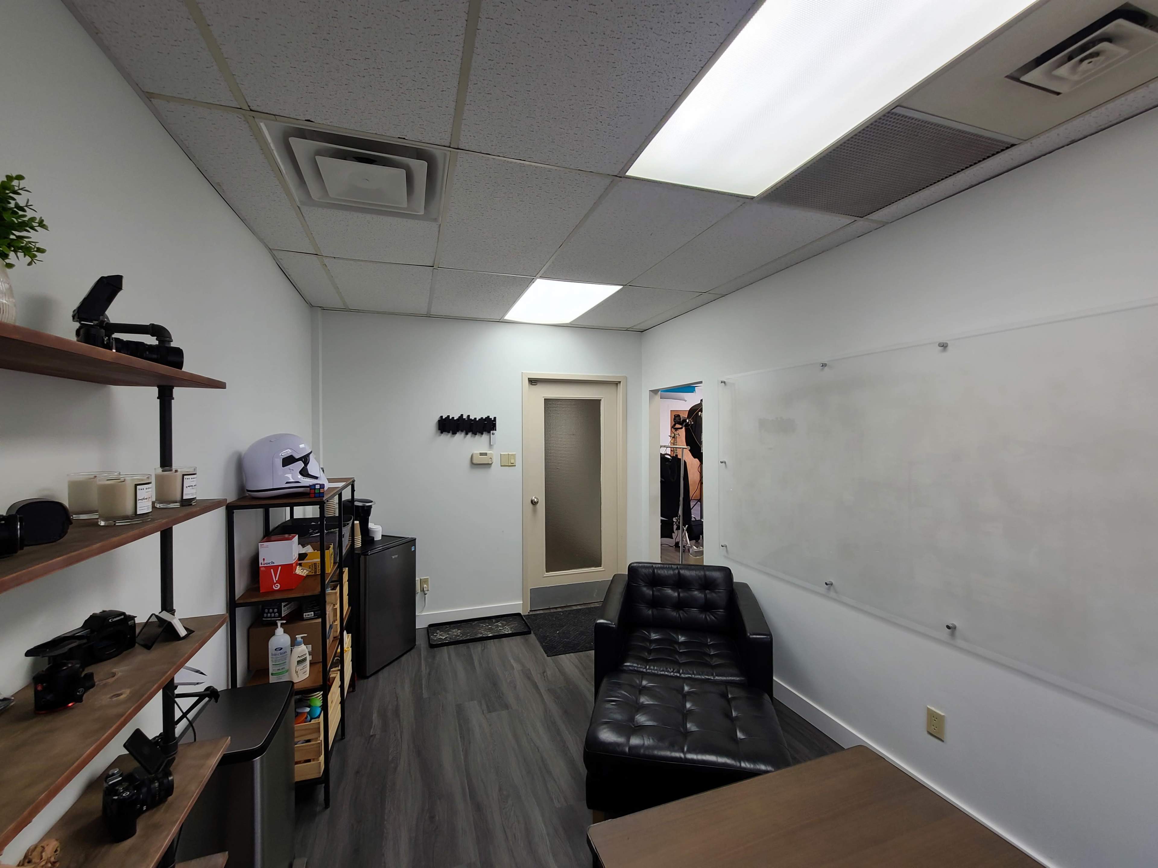 A small office space with a black couch, a shelf featuring cameras and other items, and a whiteboard on the wall.