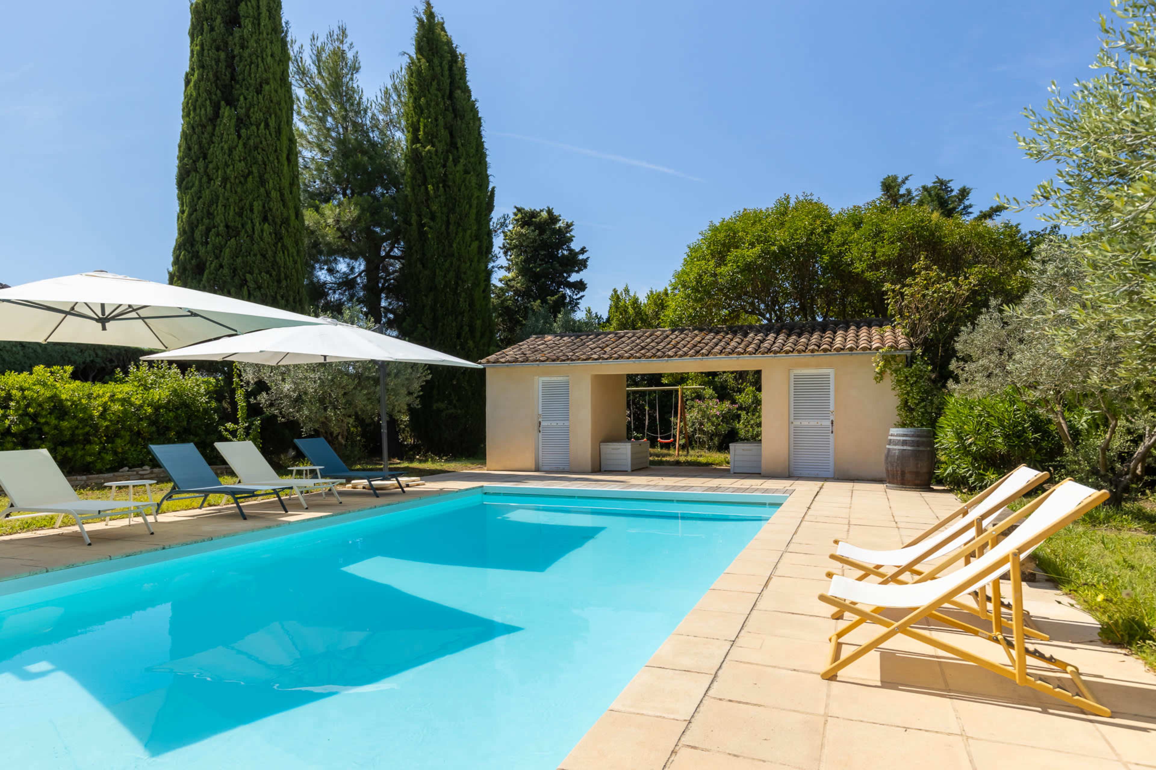 The image shows a swimming pool surrounded by lounge chairs and tall greenery under a clear sky.