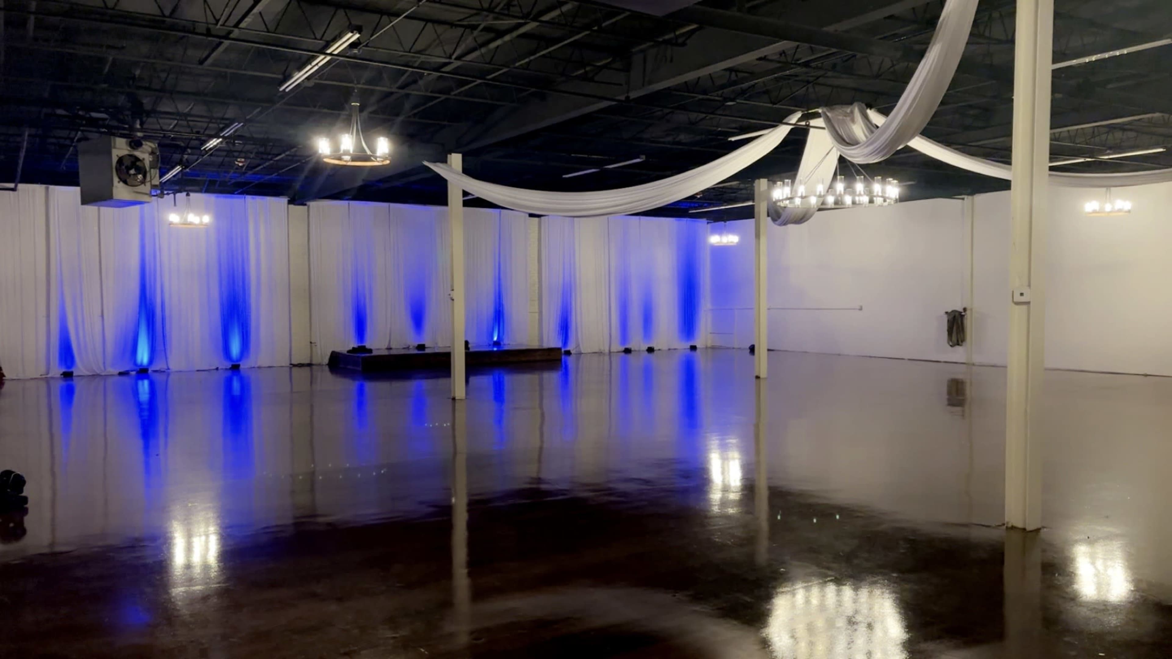 The image shows a spacious, empty event venue with blue lighting, draped fabric, and polished floors.