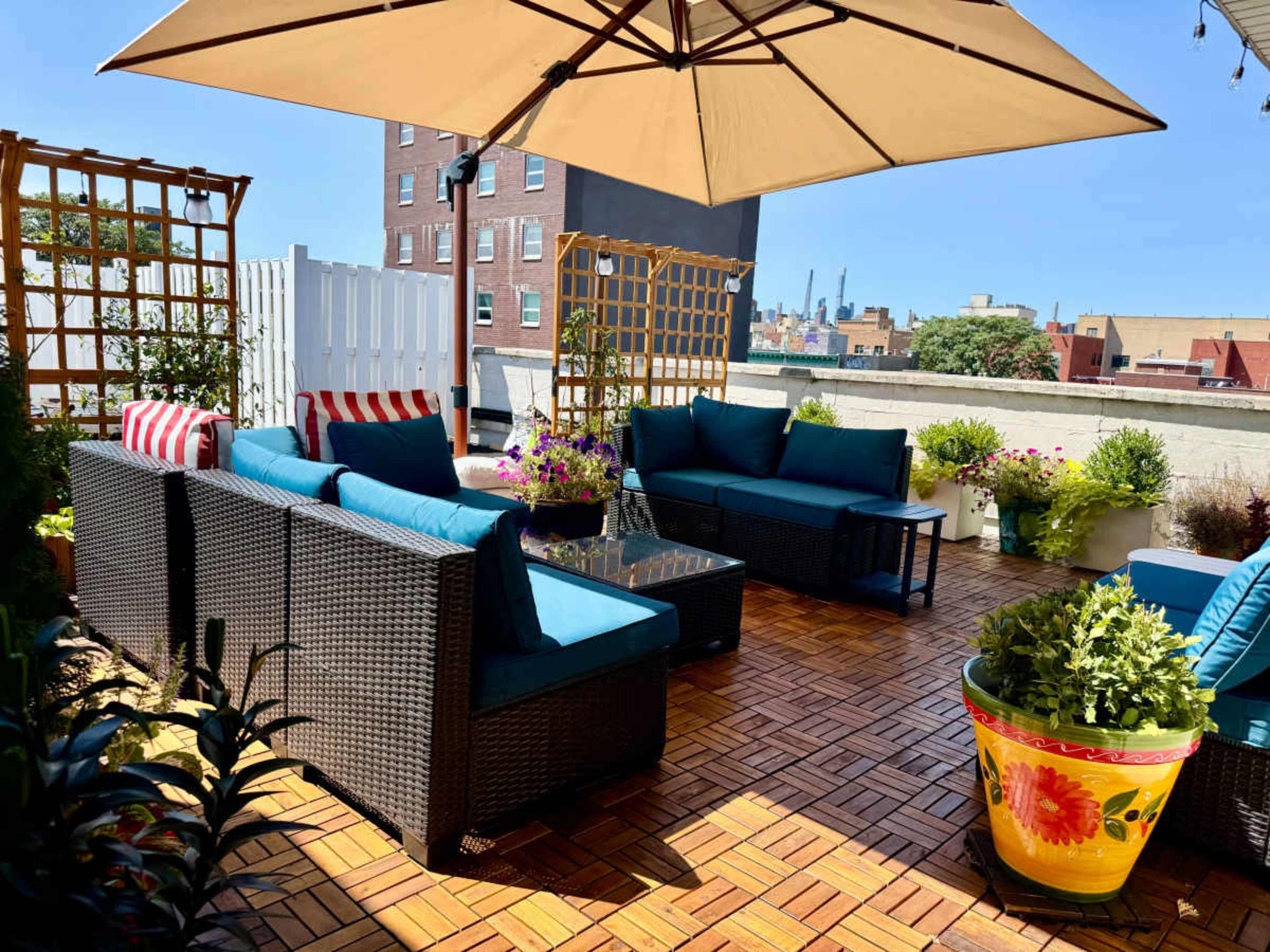 A rooftop patio features wicker furniture with blue cushions, an umbrella for shade, and vibrant potted plants.