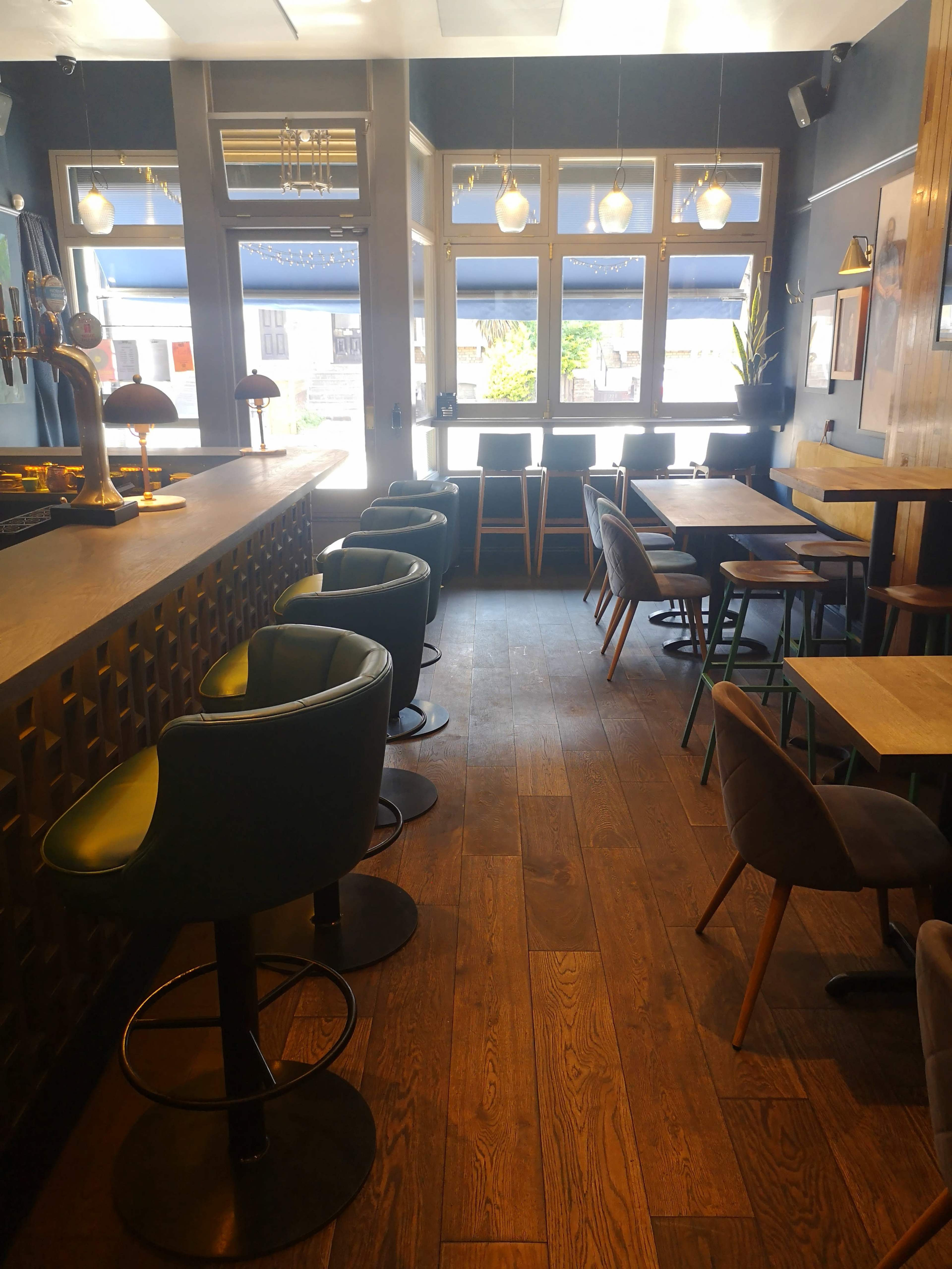 The image shows a modern bar interior with wooden flooring, high stools at the counter, and small tables arranged along the windows.