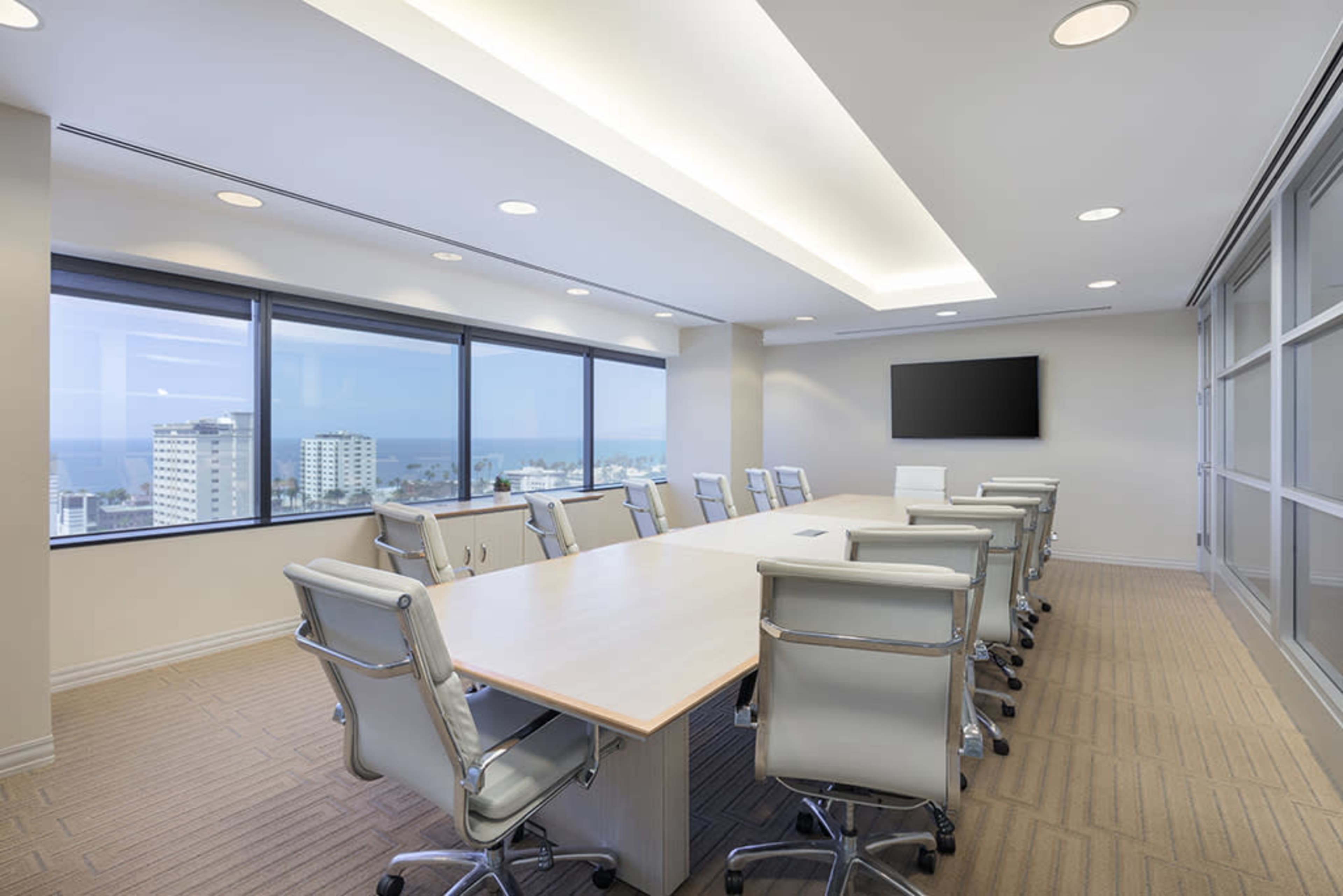 A modern conference room features a long table surrounded by ergonomic chairs, with large windows offering views of a cityscape and ocean.