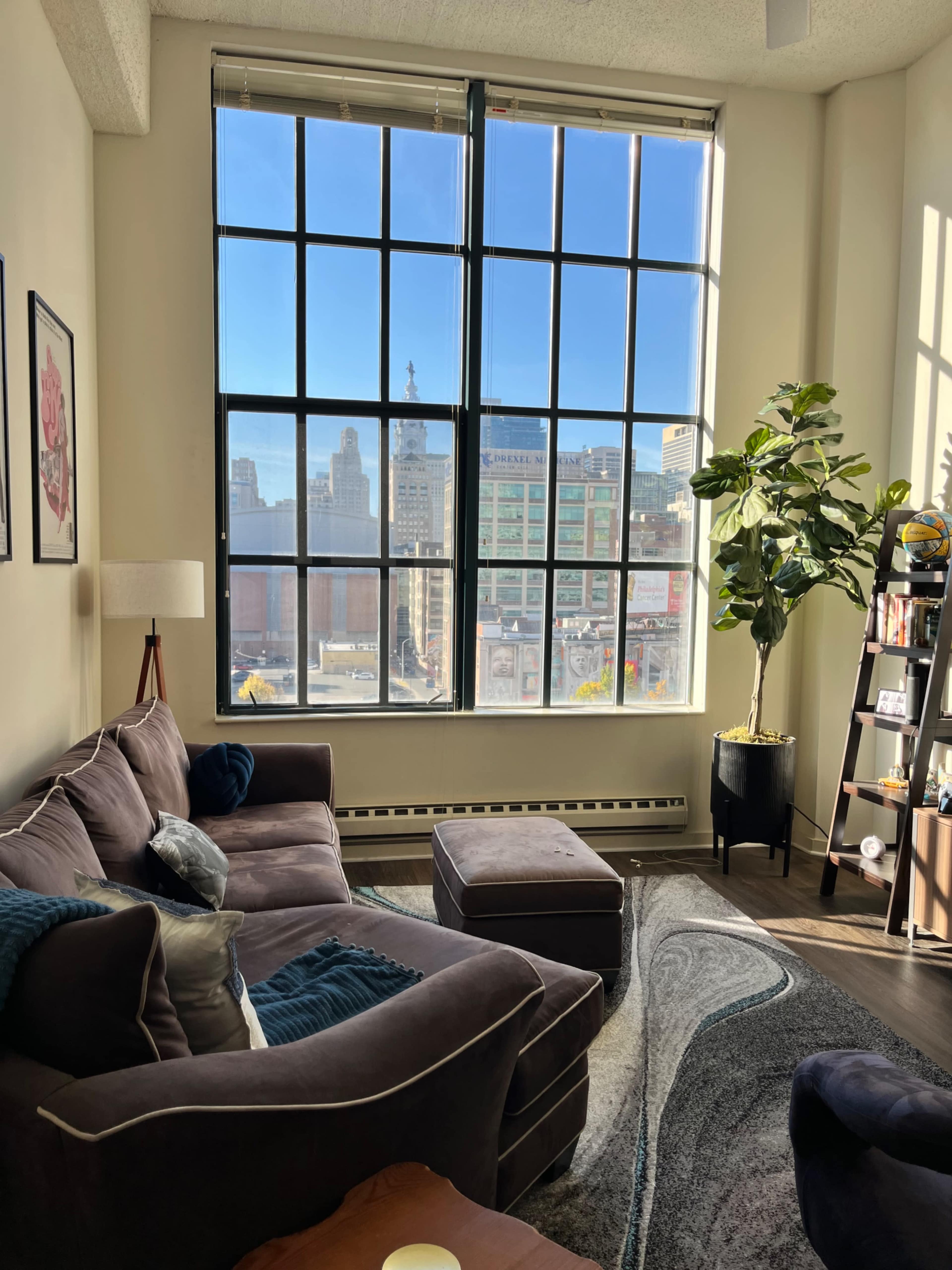 The image shows a modern living room with a large window providing a view of a city skyline, featuring a sofa, an ottoman, a potted plant, and a bookshelf.