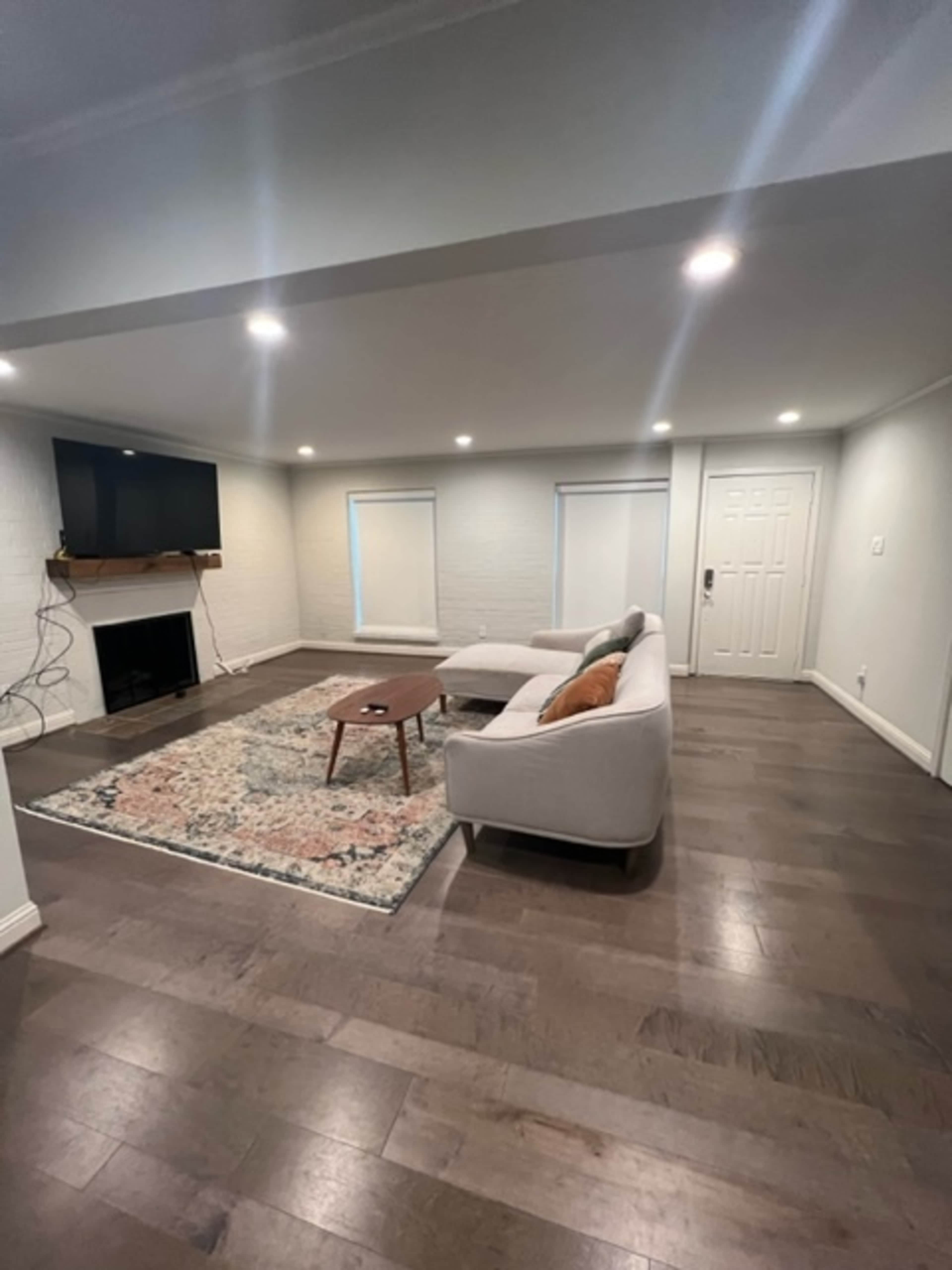A living room featuring a gray sofa, a wooden coffee table, a television mounted on a white brick wall, and two windows with closed white blinds.