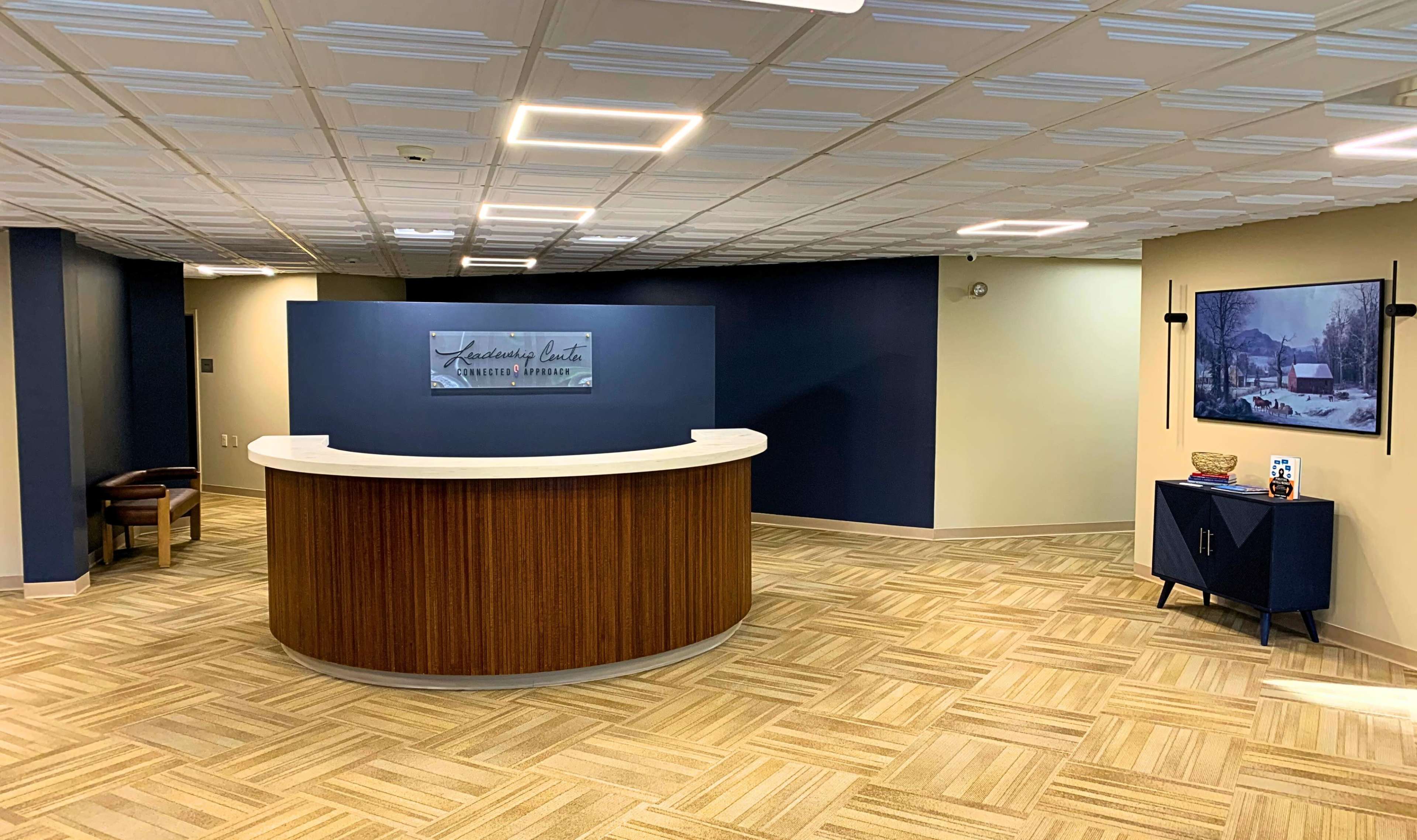The image shows a modern reception area with a curved wooden front desk, beige walls, and a framed picture on the right.