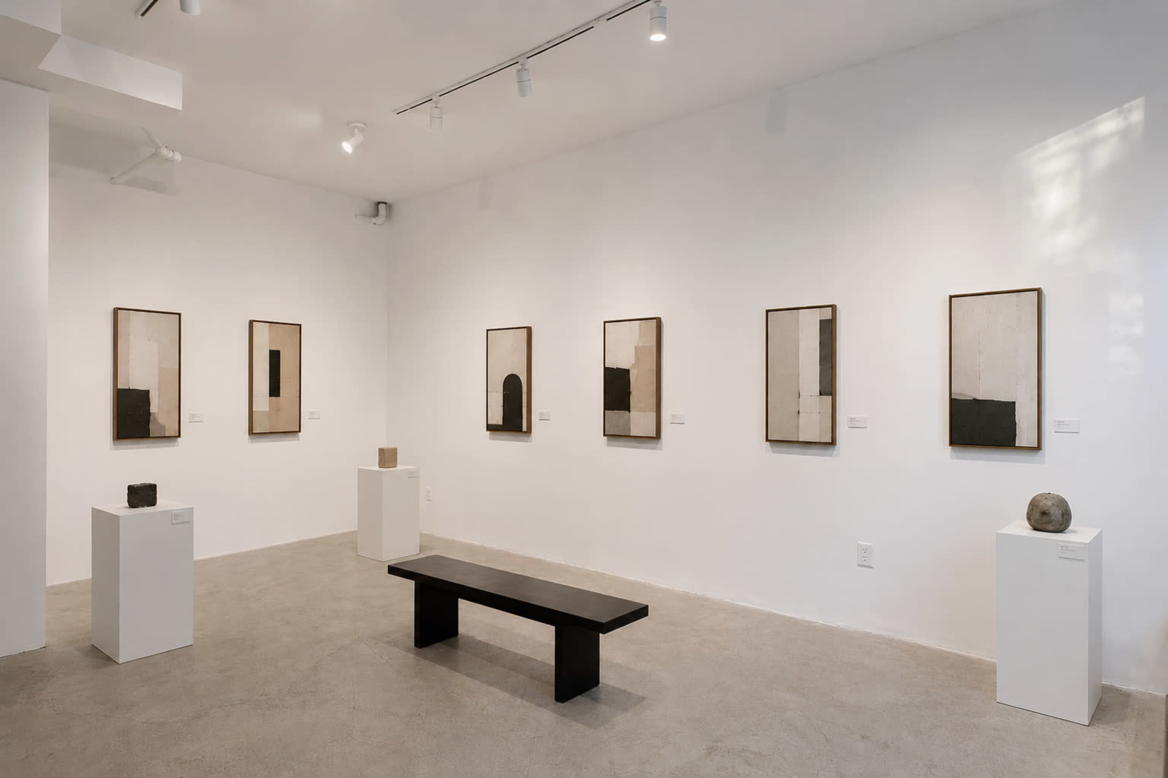 The image shows an art gallery featuring six framed abstract artworks on white walls and two small sculptures displayed on pedestals.