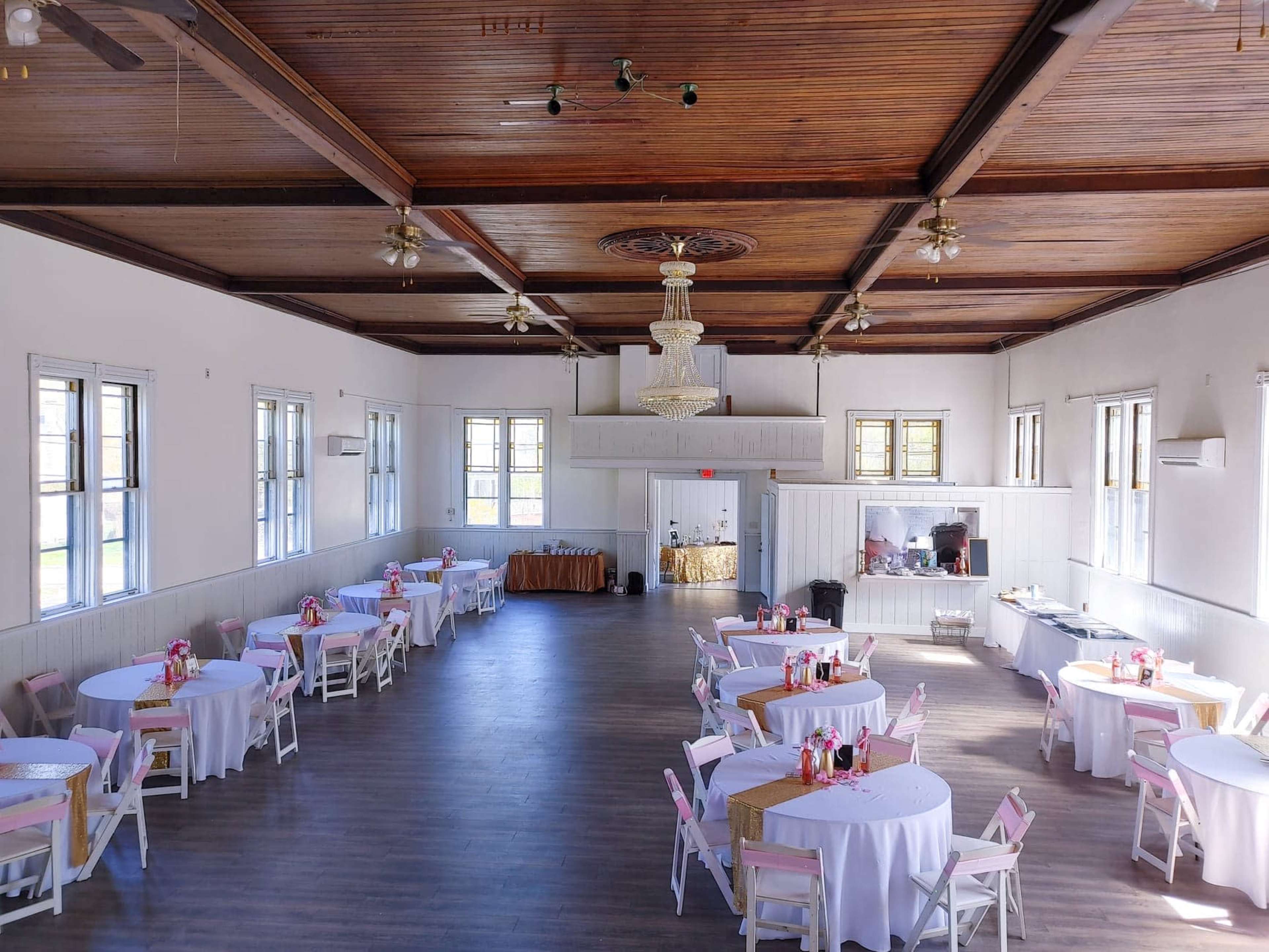 The image shows a spacious hall with round tables covered in white tablecloths and pink accents, arranged calmly around the room beneath a chandelier and wooden ceiling.